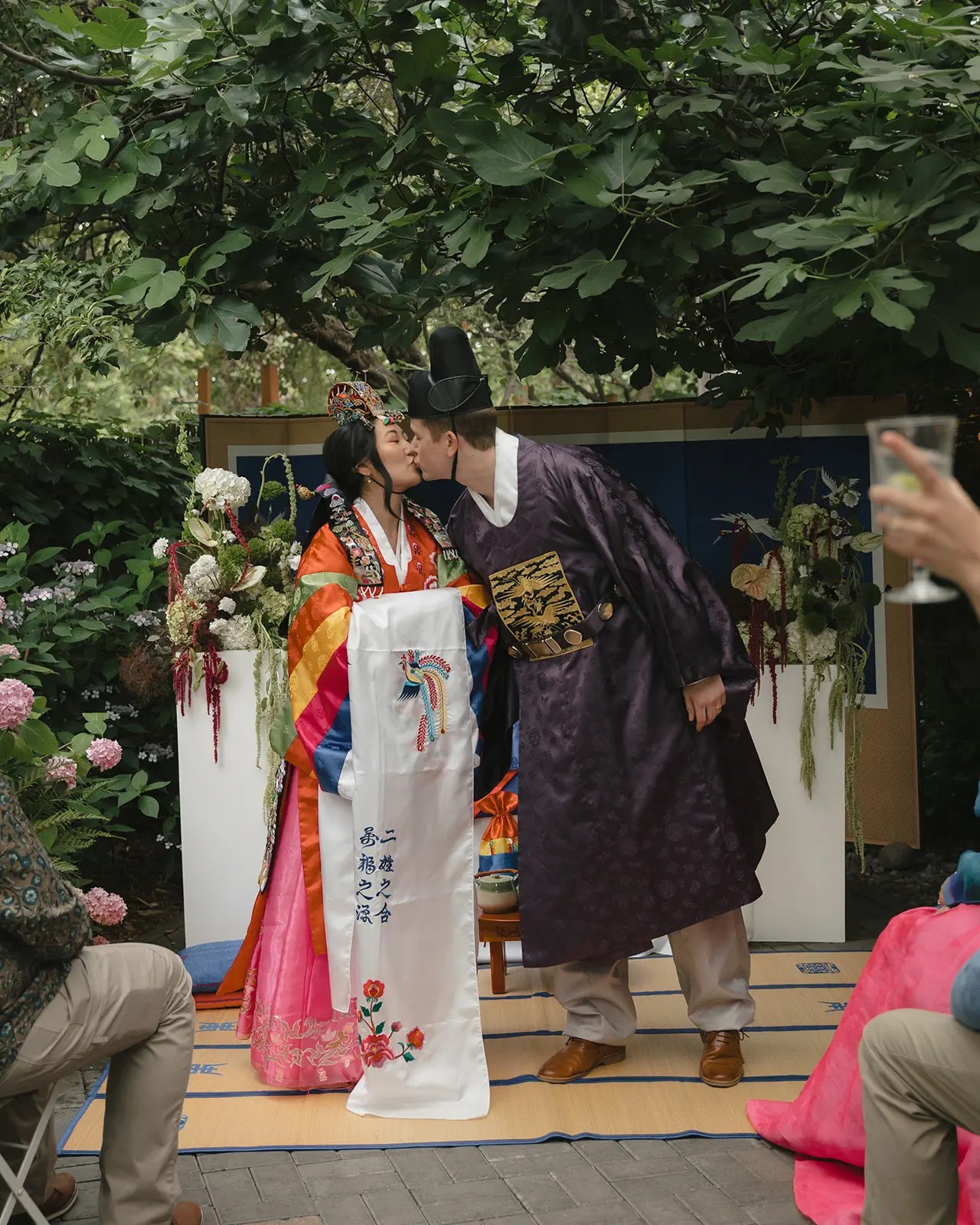 Mariée en hanbok multicolore superposé et coiffe de mariée brodée embrassant le marié en robe de brocart violet et chapeau noir lors d'une cérémonie Paebaek coréenne en plein air avec écran de grue bleu