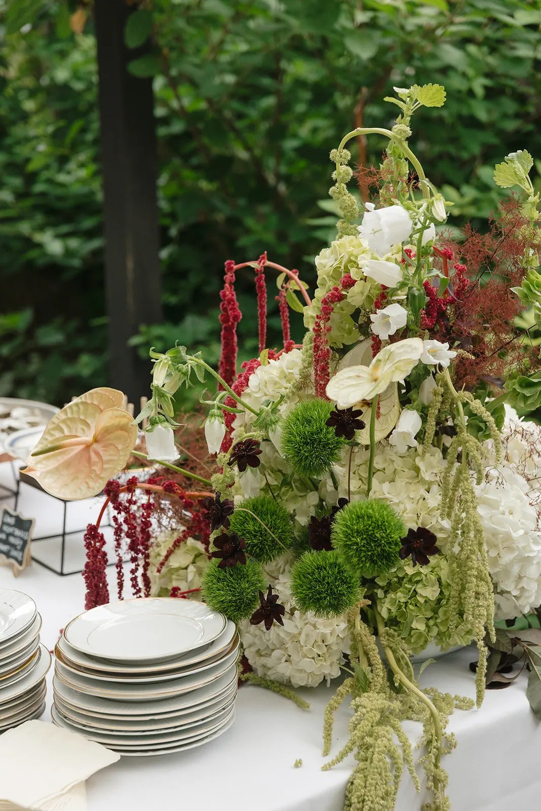 Centre de table de mariage dans le jardin composé d'hortensia blanc, d'amarante pourpre, de fleurs à pompons verts et d'anthurium crème à côté d'assiettes empilées cerclées d'or