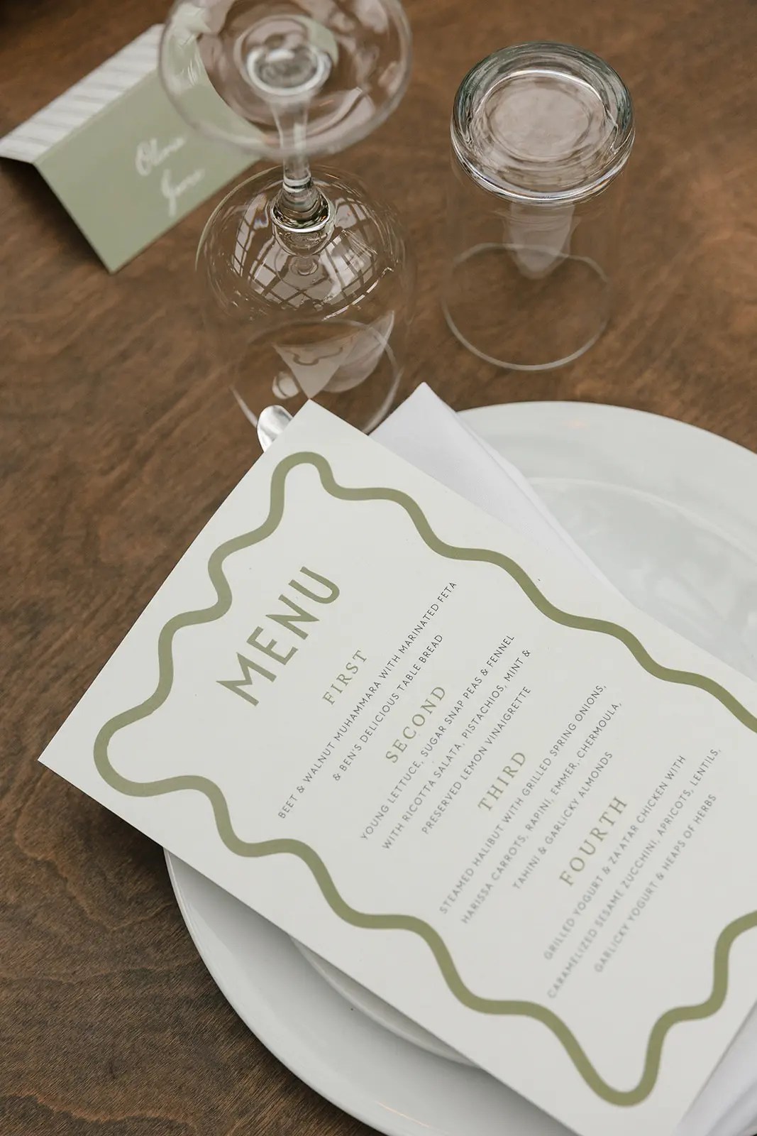 Carte de menu de mariage à quatre plats avec bordure ondulée vert sauge sur une assiette blanche sur une table en bois sombre