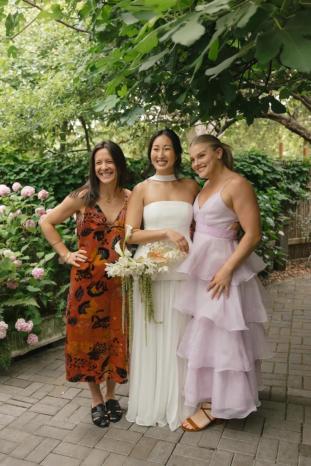 Mariée en robe blanche sans bretelles et tour de cou blanc tenant un bouquet d'orchidées blanches entre amis en imprimé botanique rouille et robes en organza lavande à plusieurs niveaux sur une allée de jardin