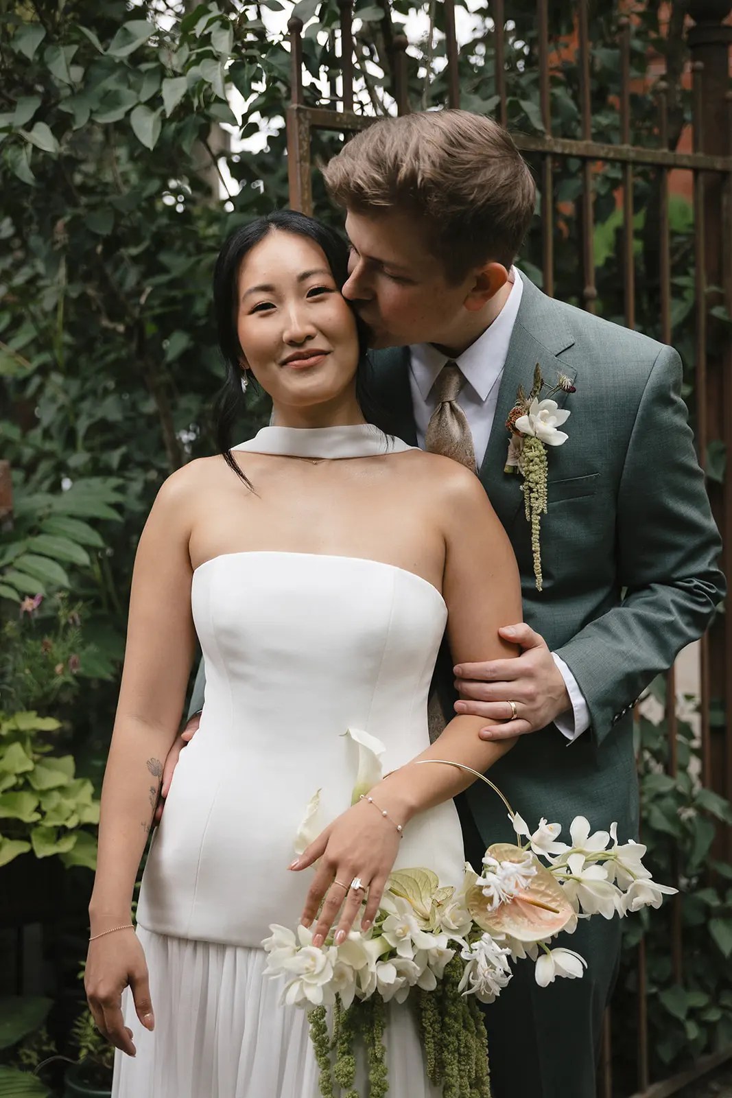 Marié en costume vert sauge embrassant la joue de la mariée par derrière, mariée en robe blanche dos nu avec bouquet de cerceaux devant une porte de jardin en fer recouverte de vigne