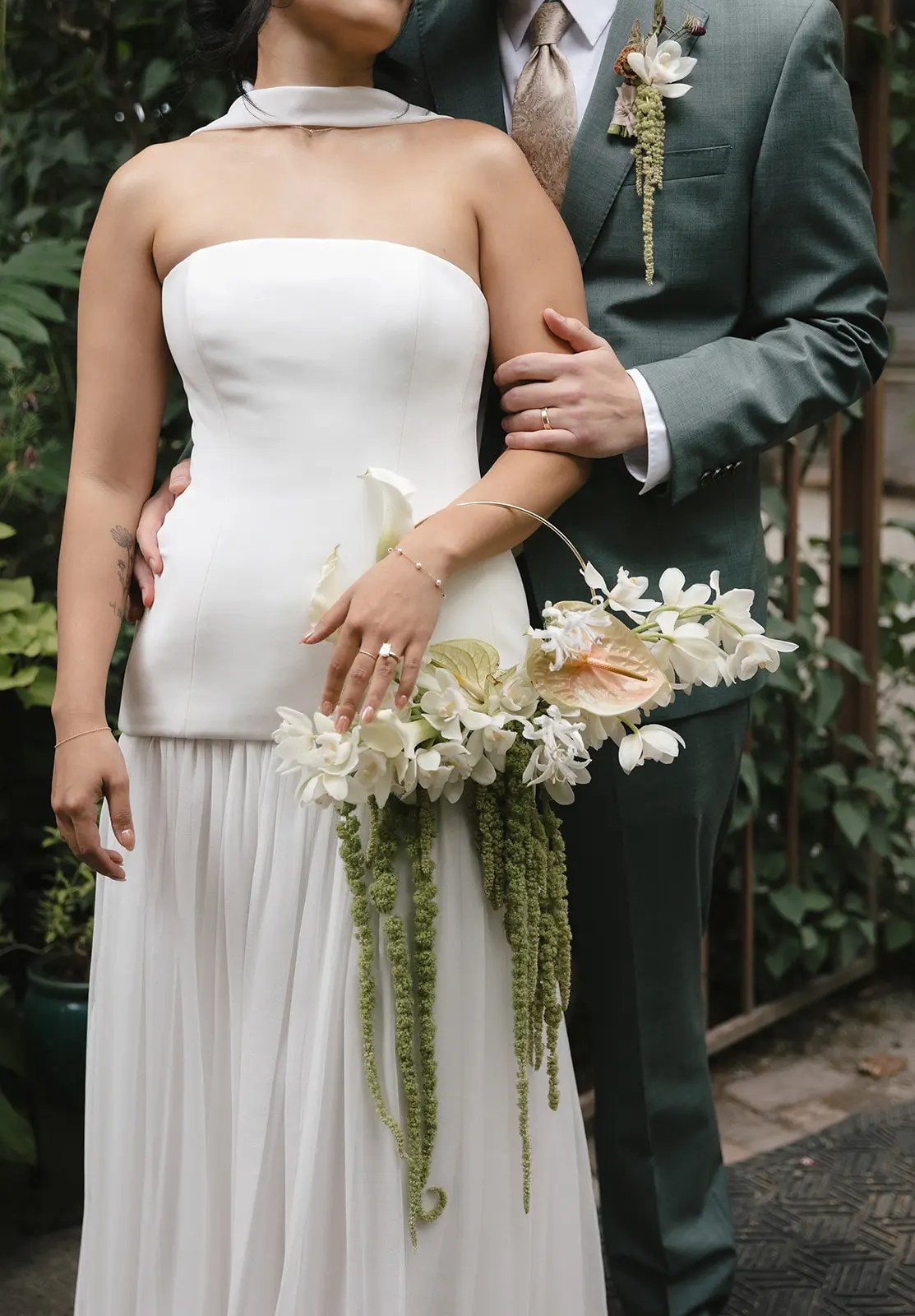 Détail d'une robe de mariée blanche sans bretelles à taille basse, d'une manche de costume vert sauge avec une alliance dorée et d'un bouquet d'orchidées blanches et d'amarante verte