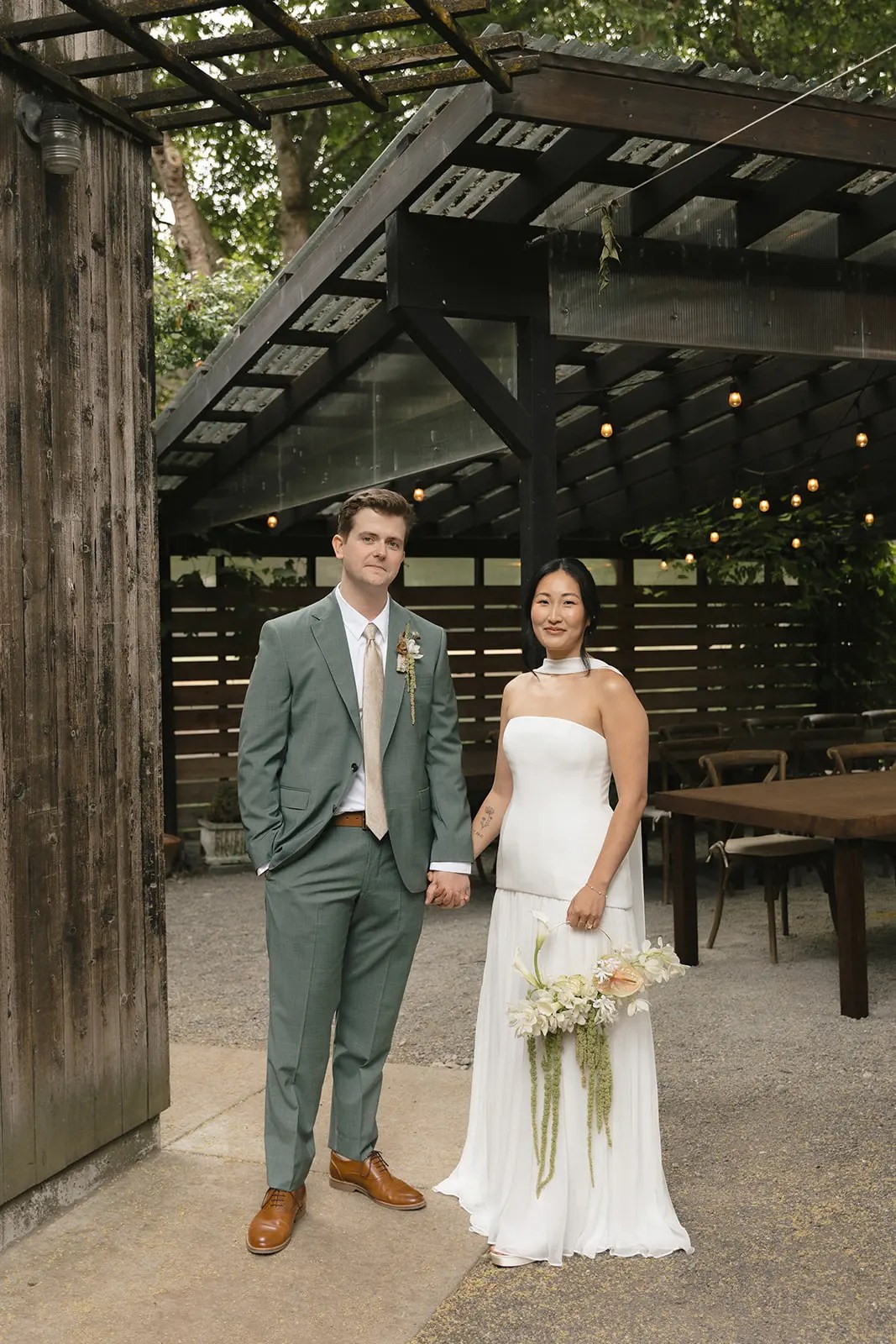 Tess and Connor holding hands under a string-lit pergola at The Corson Building in Seattle, groom in sage green Jack Victor suit, bride in white strapless Alexandra Grecco gown with hoop bouquet