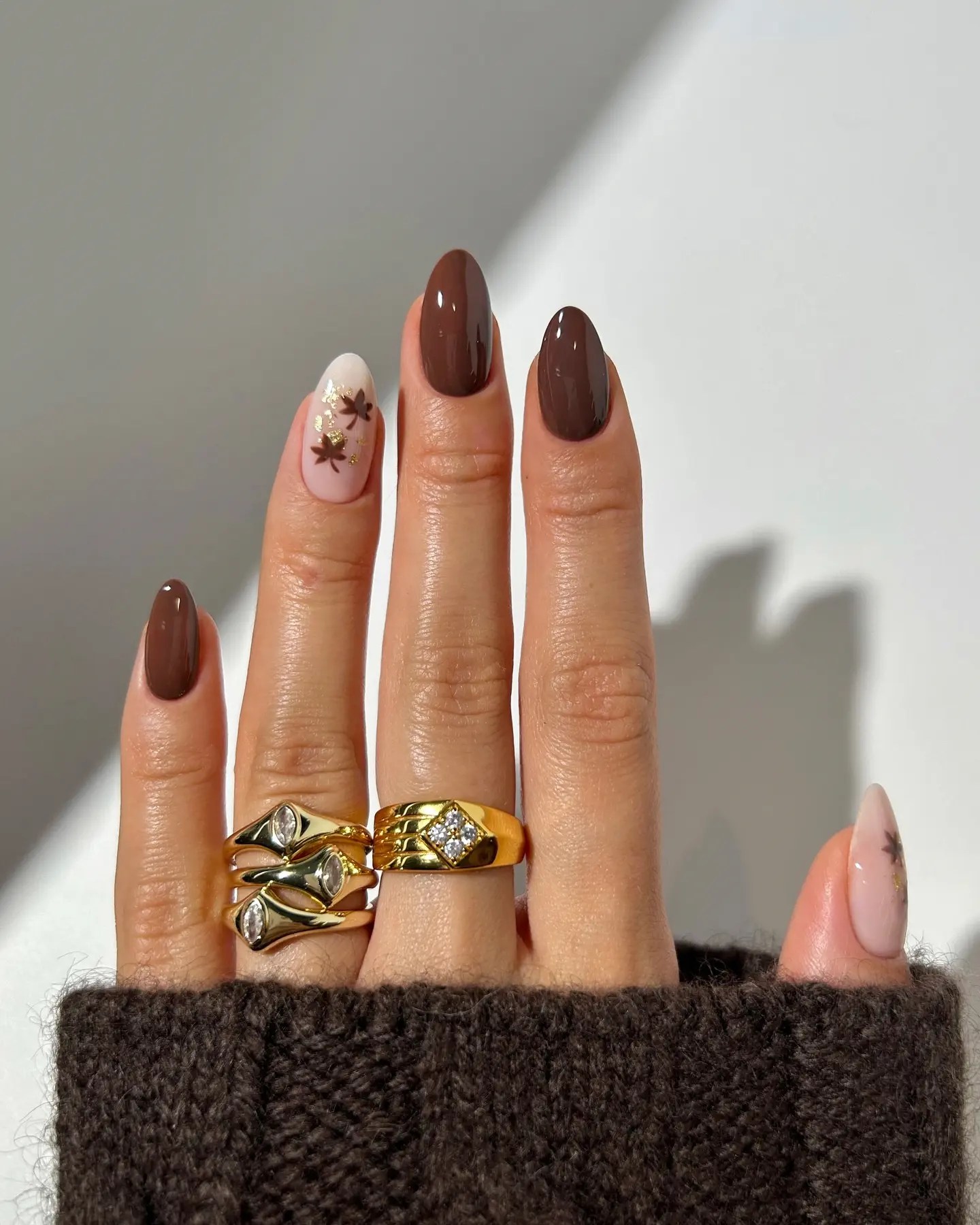 Chocolate brown almond nails with one milky white accent nail featuring small brown maple leaves and gold foil flakes.