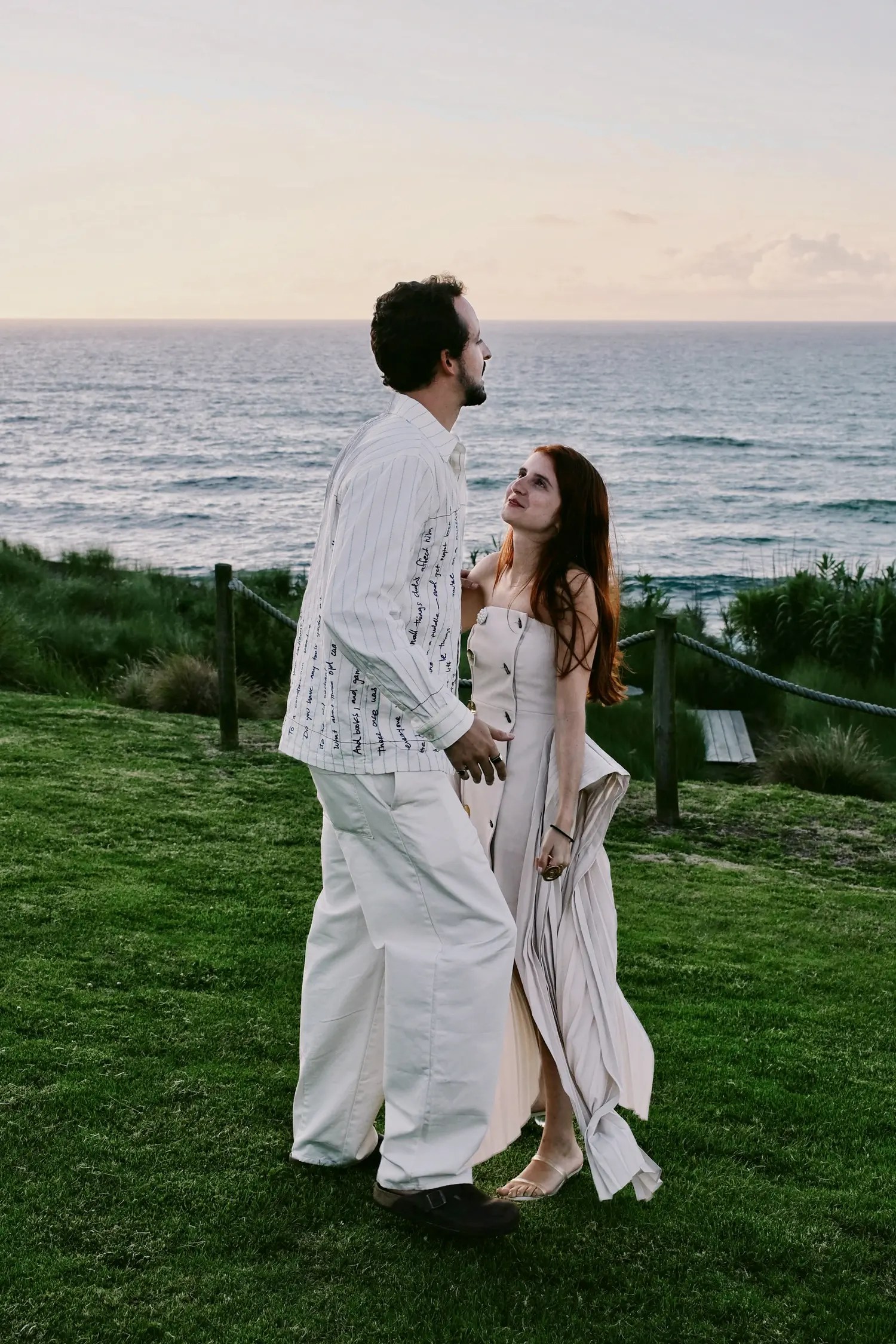 Couple at sunset on a clifftop lawn at Santa Barbara Eco Beach Resort, groom in KidSuper Studios handwriting-embroidered overshirt, bride in customized cream ASOS button-front maxi dress
