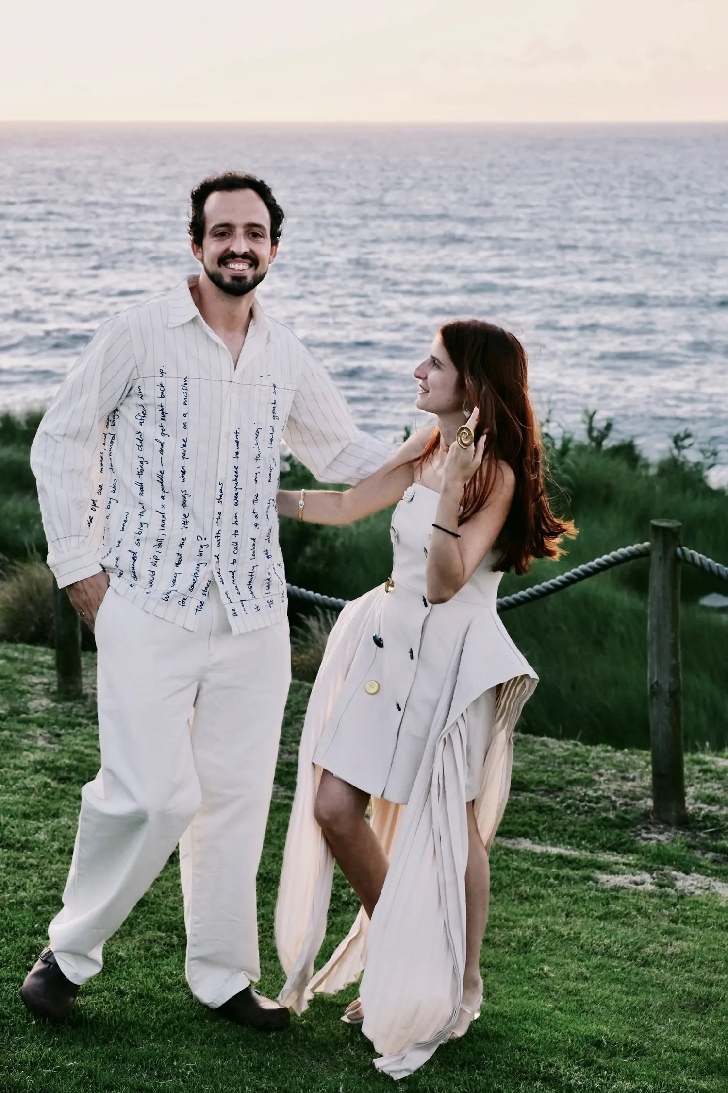 Couple laughing at an ocean-view welcome dinner in the Azores, bride in cream strapless dress with oversized buttons and gold spiral earring, groom in cursive-print shirt and cream trousers