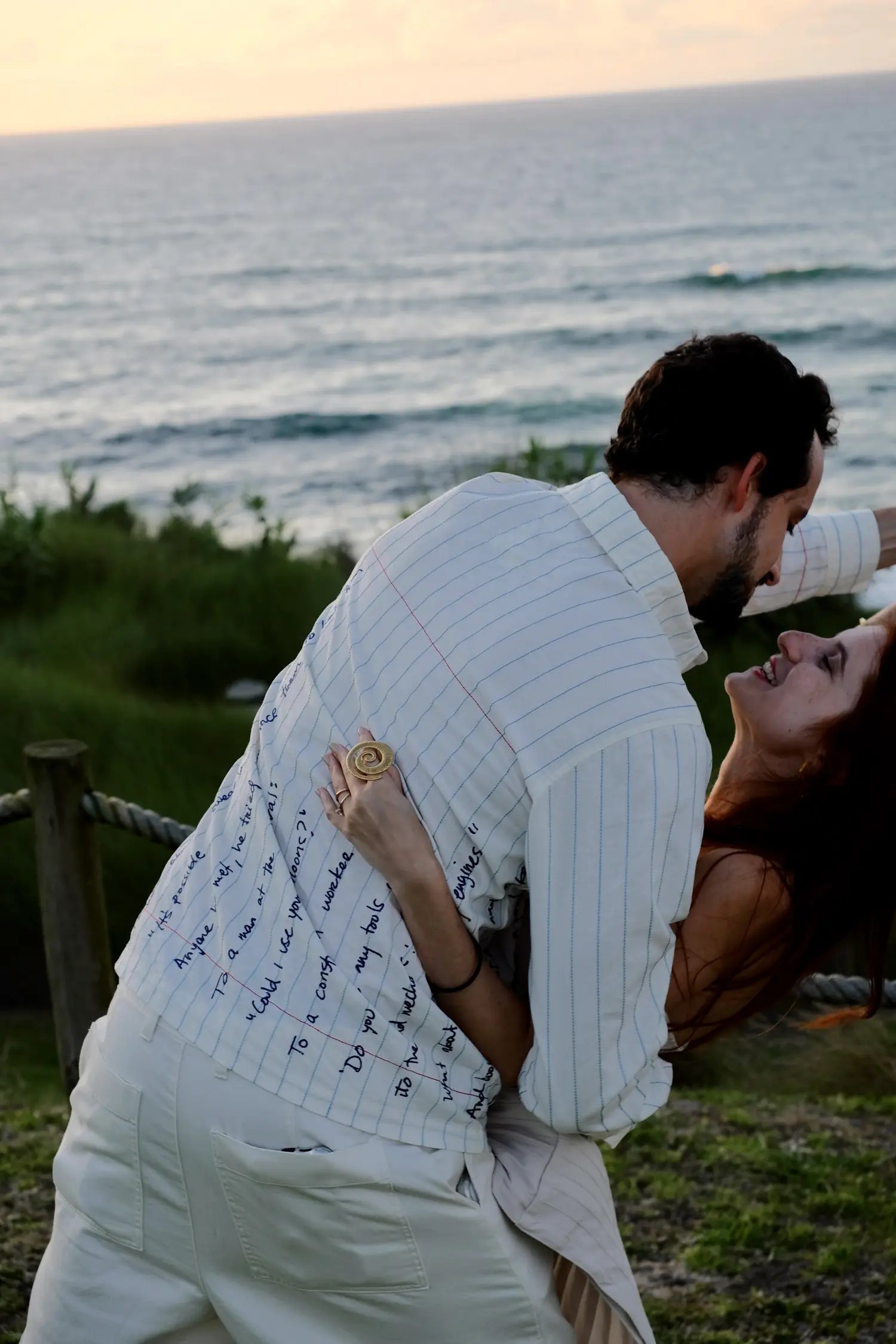 Bride laughing and leaning back as groom dips her at a clifftop welcome dinner, his navy script-print white shirt filling the frame, gold coil ring on her hand