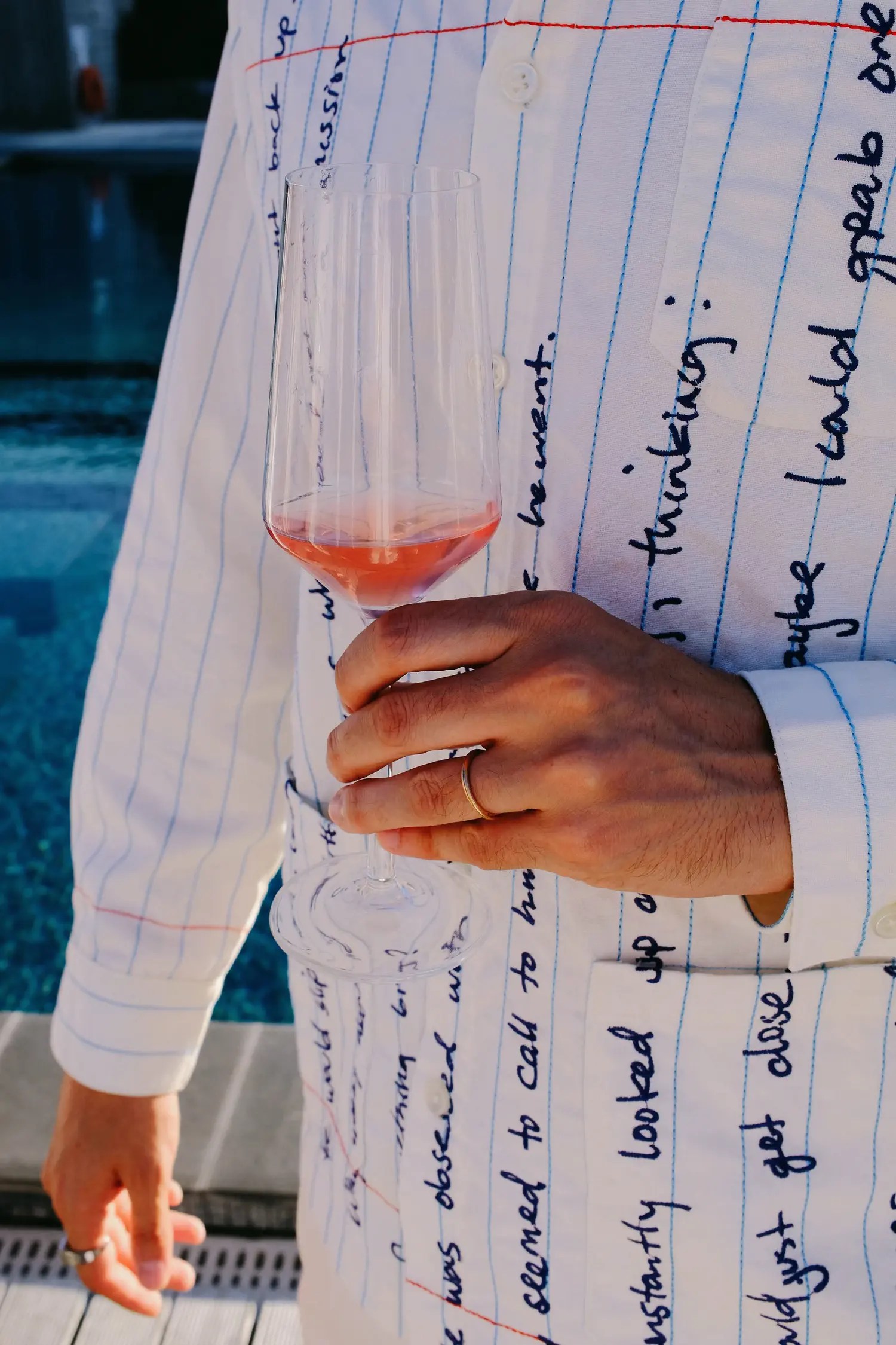 Hand with wedding band holding a rosé champagne flute against a notebook-print white KidSuper Studios shirt, swimming pool blurred behind