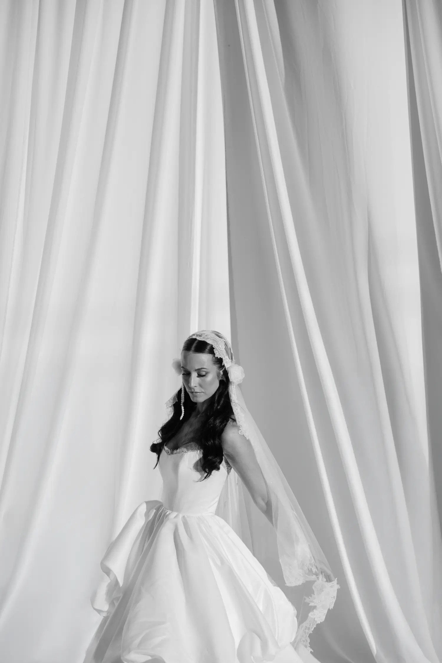 Portrait noir et blanc d'une mariée vêtue d'une robe de bal blanche sans bretelles avec un voile de dentelle Juliette et de longues boucles d'oreilles en chaîne de perles contre des rideaux blancs swagged