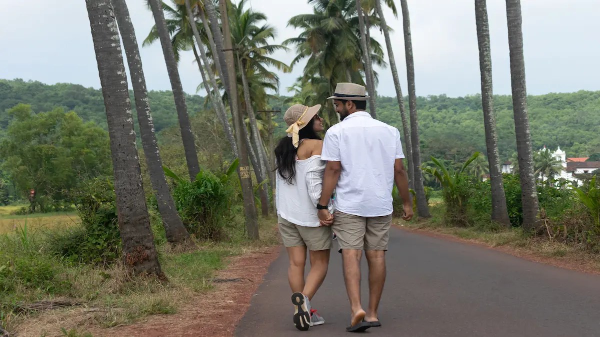 Couple walking hand-in-hand down a palm-lined road during the Goa monsoon, surrounded by lush greenery and overcast skies.