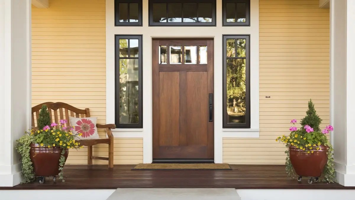 A welcoming front porch featuring a wooden door with glass panels, flanked by tall black-framed windows. Two large planters filled with colorful flowers sit on either side of the entrance, and a wooden bench with a floral cushion is placed to the left against the yellow siding wall.