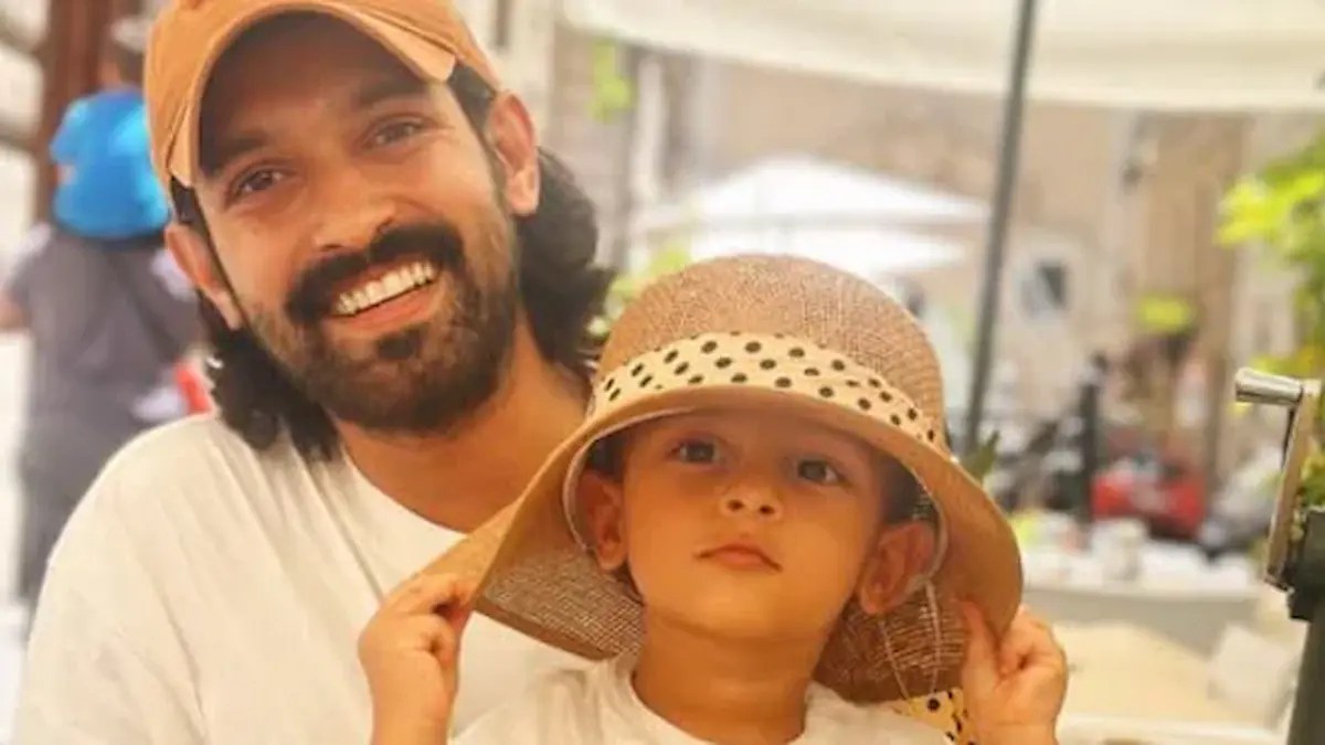 A smiling man sitting with a small child wearing a hat, sharing a cheerful moment together.