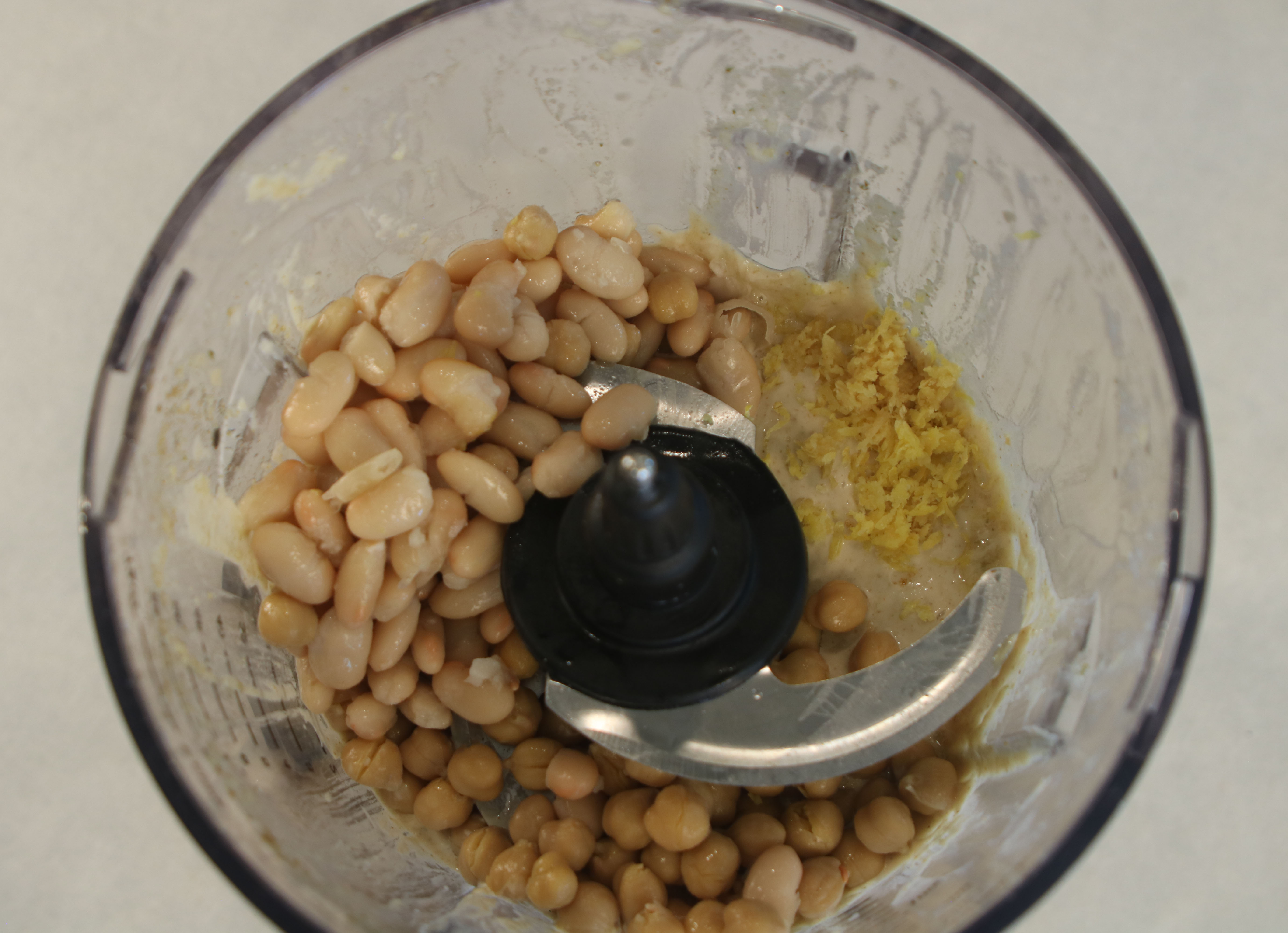 An overhead view of a food processor bowl filled with chickpeas, white beans, and lemon zest, ready to be blended into hummus.