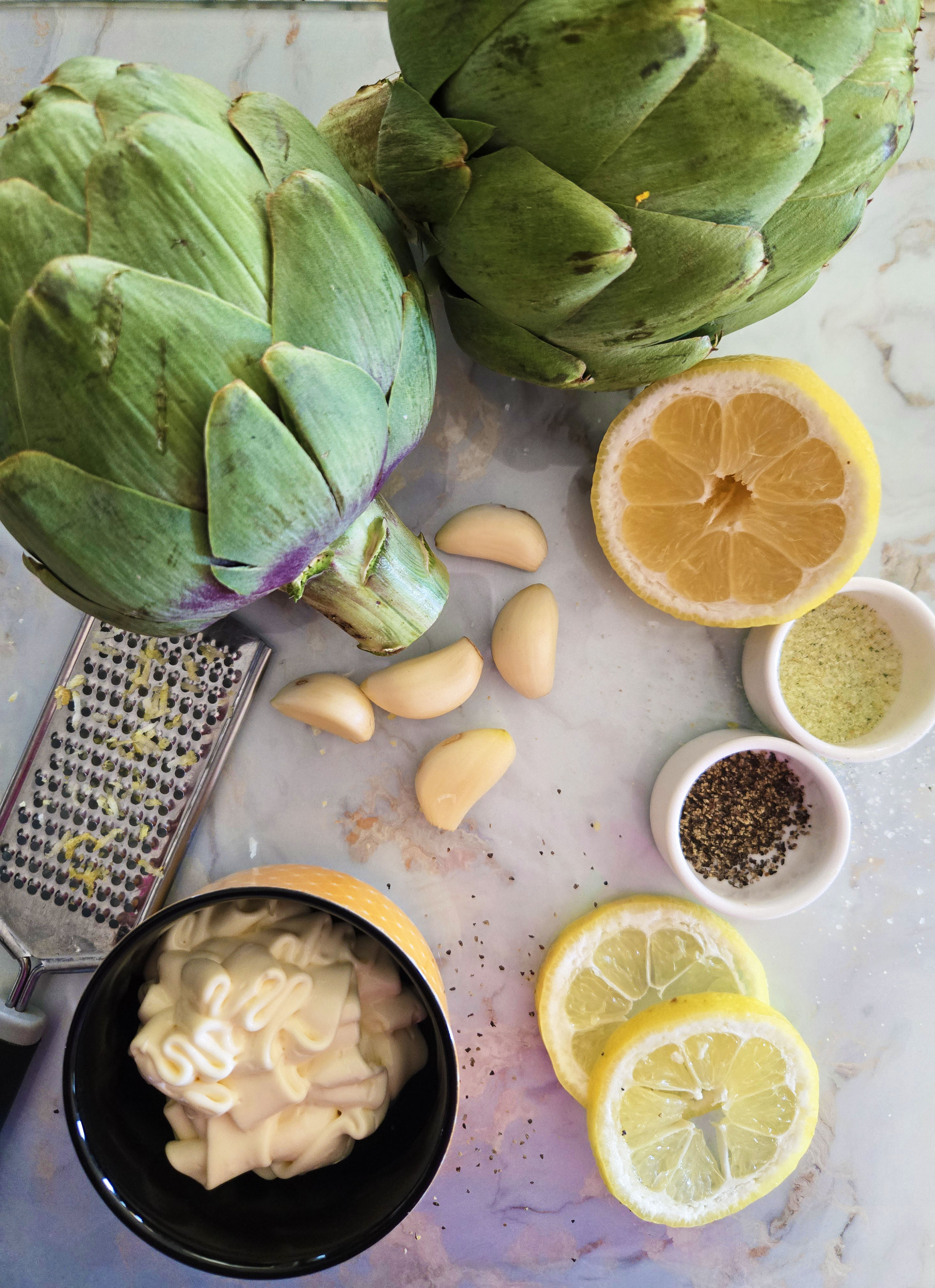Fresh artichokes with garlic cloves, lemon slices, mayonnaise, and seasonings arranged on a marble surface—ingredients for lemon garlic aioli.
