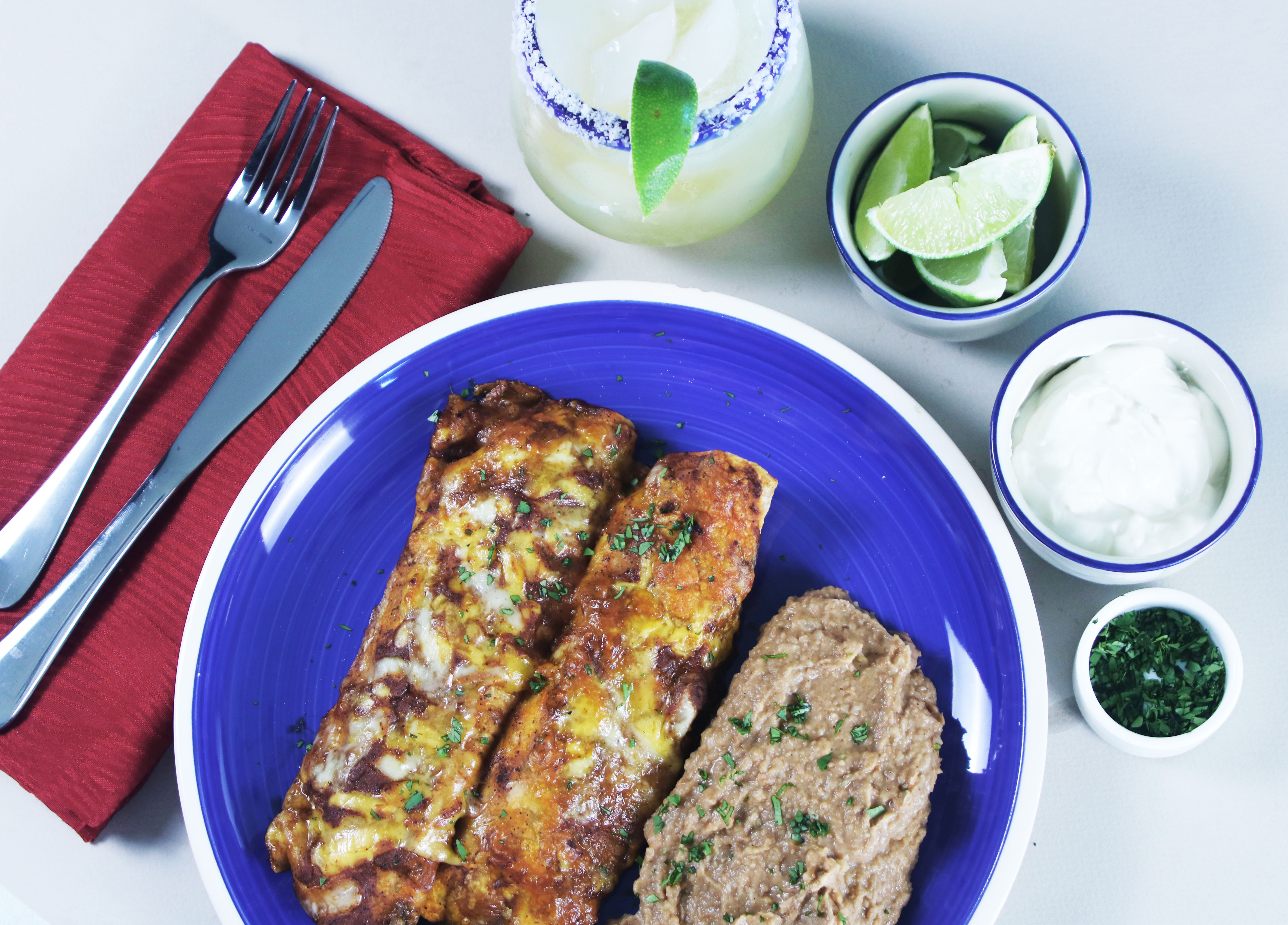 Two cheesy supreme beef enchiladas topped with red enchilada sauce and served with creamy refried beans on a vibrant blue plate, paired with lime wedges, sour cream, and a margarita.