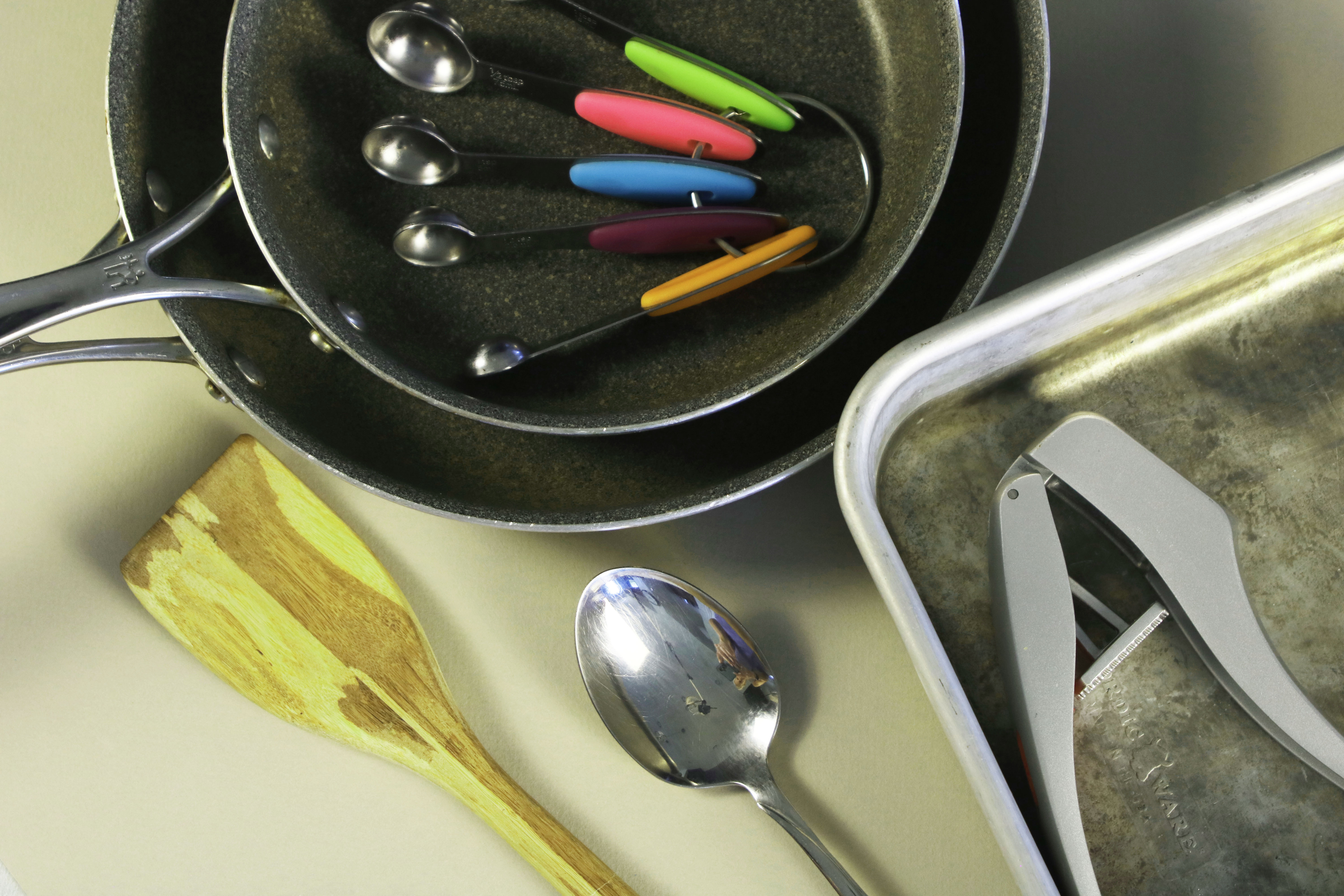 Overhead shot of assorted cooking tools for beef enchiladas, including stacked nonstick pans, colorful measuring spoons, a wooden spatula, metal spoon, garlic press, and a worn sheet pan on a neutral surface.