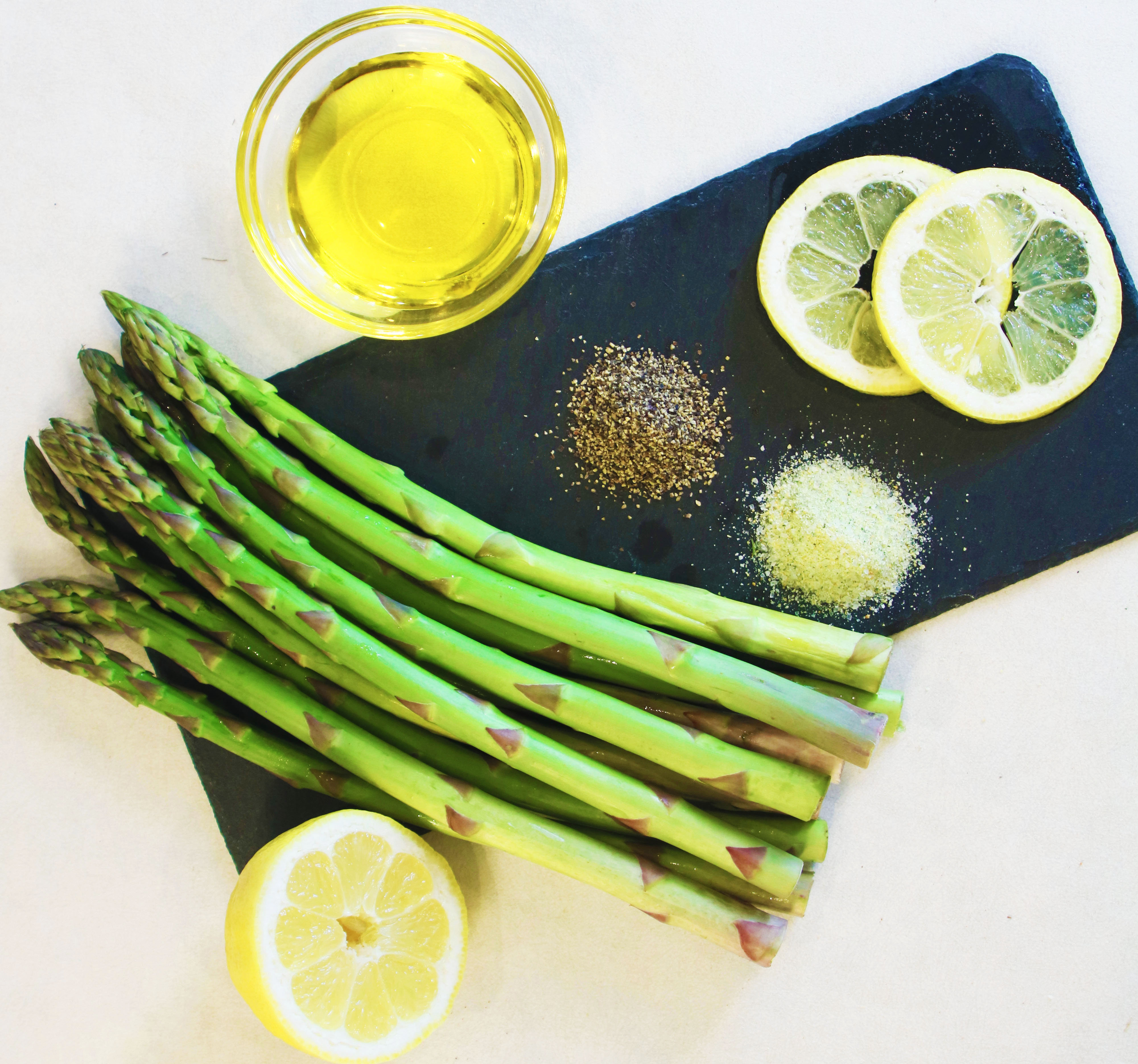 Fresh asparagus spears with half a lemon, lemon slices, a small glass bowl of olive oil, black pepper, and lemon pepper seasoning on a black slate board.