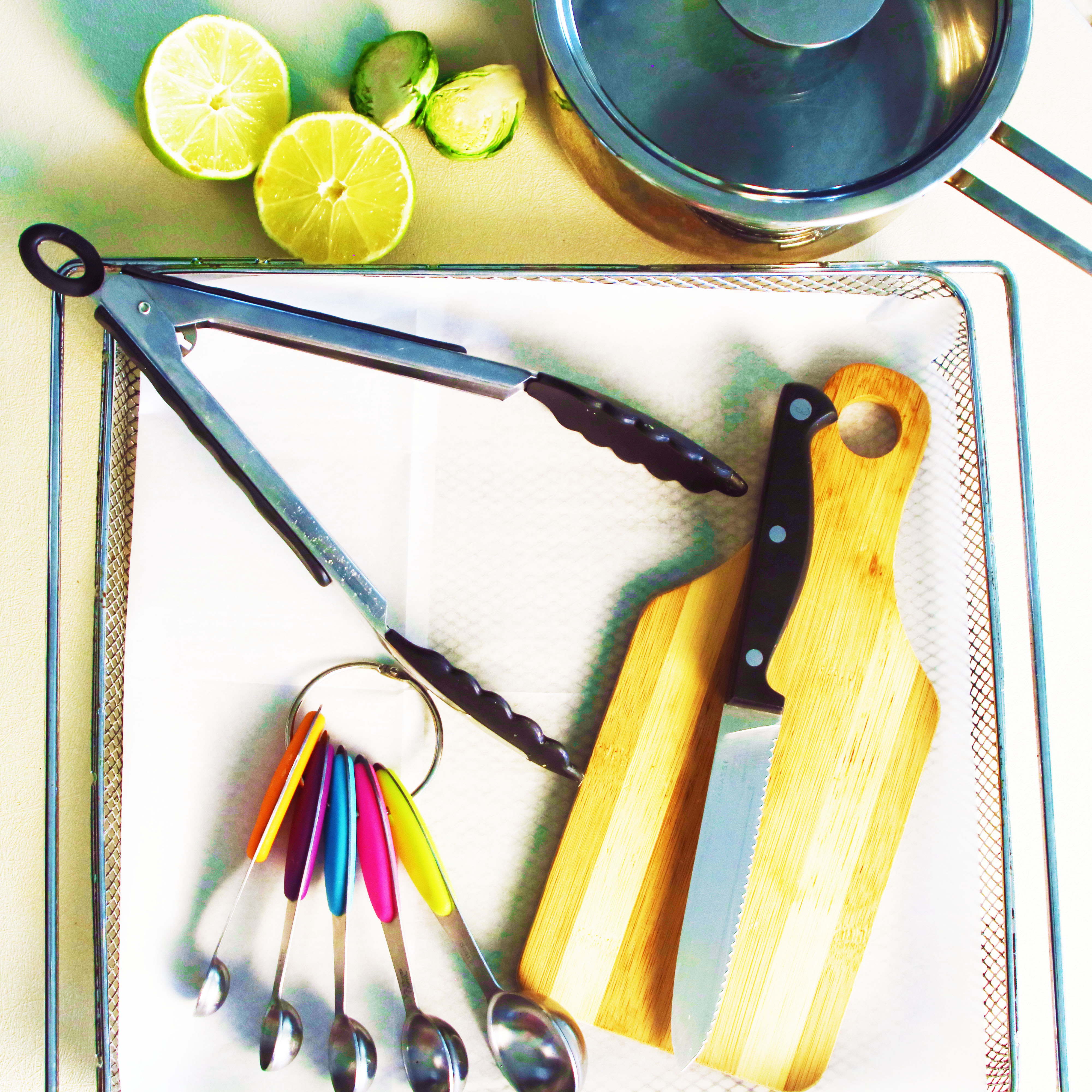Air fryer Brussels sprouts prep tools including tongs, cutting board with knife, saucepan, and measuring spoons arranged with fresh lime and halved sprouts.