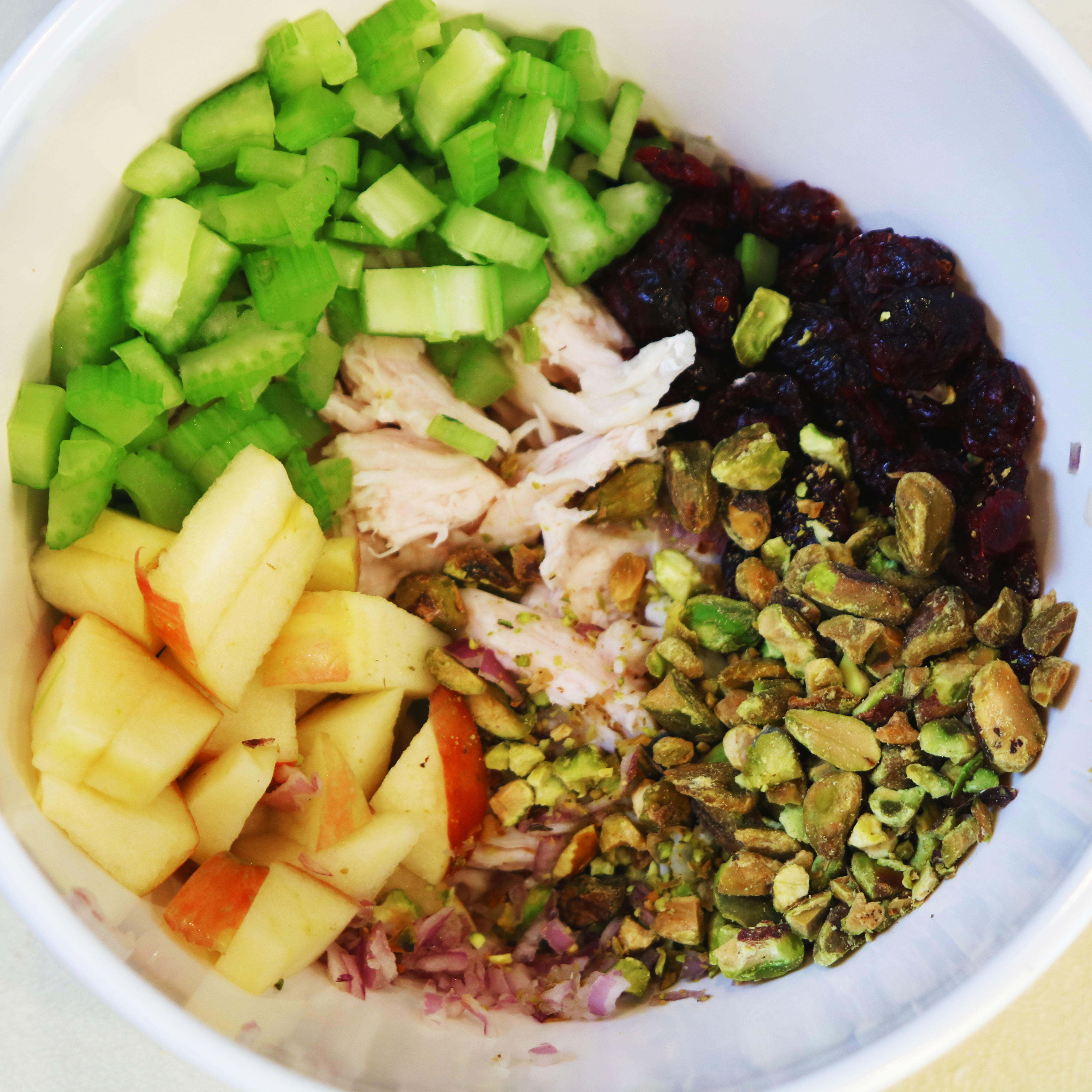 Chopped apples, celery, pistachios, dried cranberries, red onion, and shredded chicken in a bowl before mixing into salad.