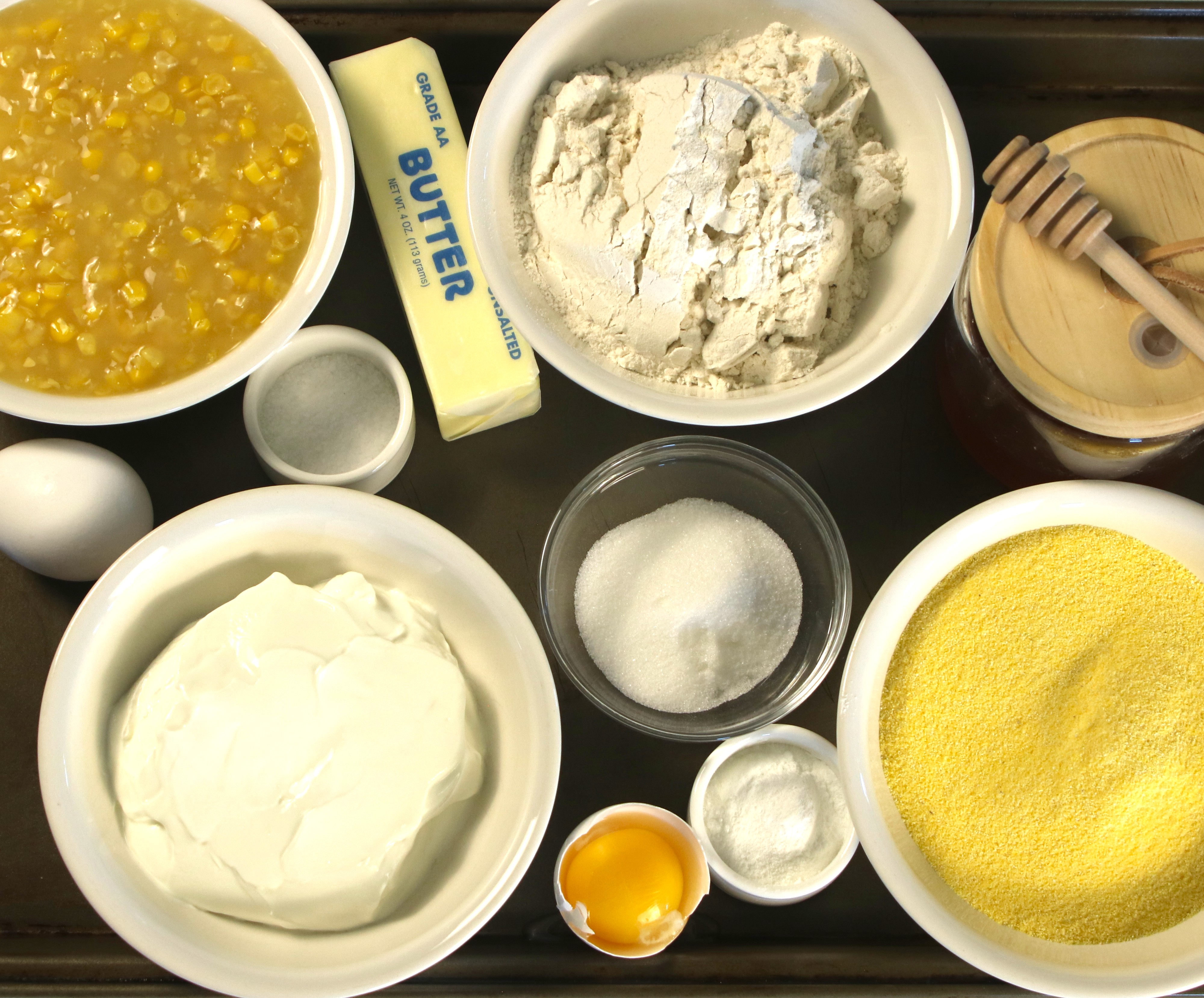 Overhead of ingredients for sweet cream cornbread—cornmeal, flour, sugar, salt, baking powder, sour cream, egg, butter, honey, and cream-style corn.
