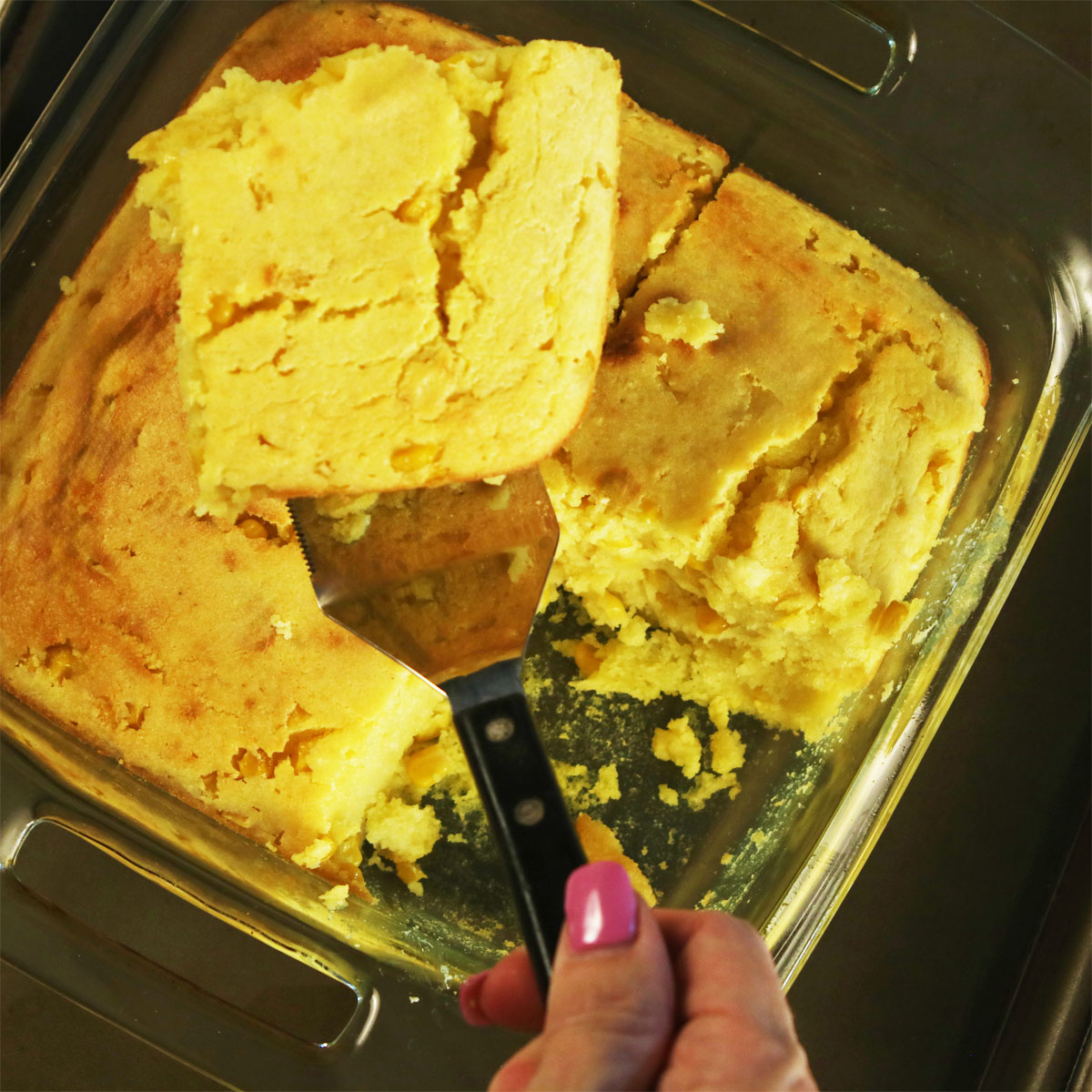 Square of sweet cream cornbread lifted from an 8×8 glass pan on a spatula, showing a tender, moist crumb.