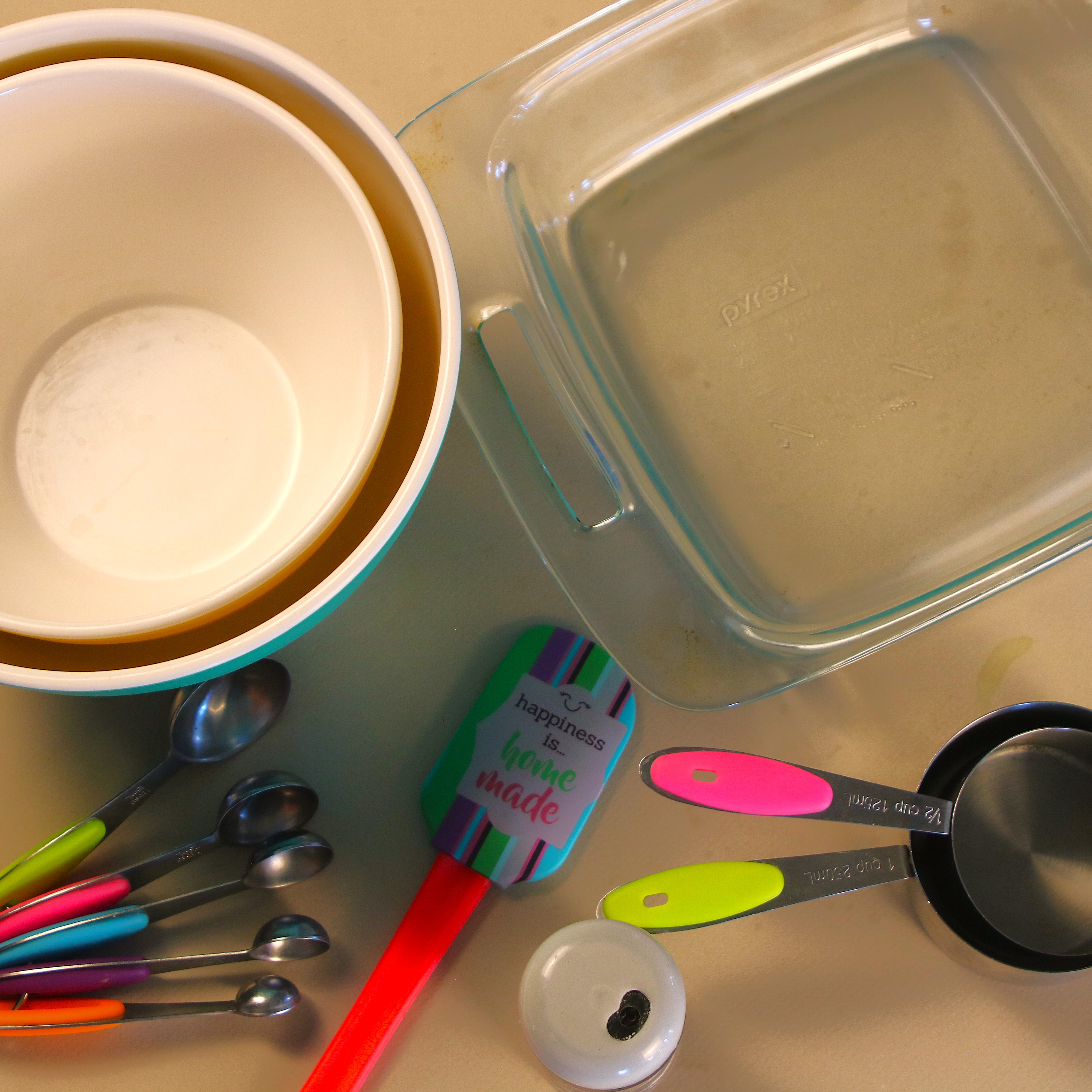 Mixing bowls, an 8×8 glass baking dish, measuring cups and spoons, and a colorful spatula arranged for baking prep.