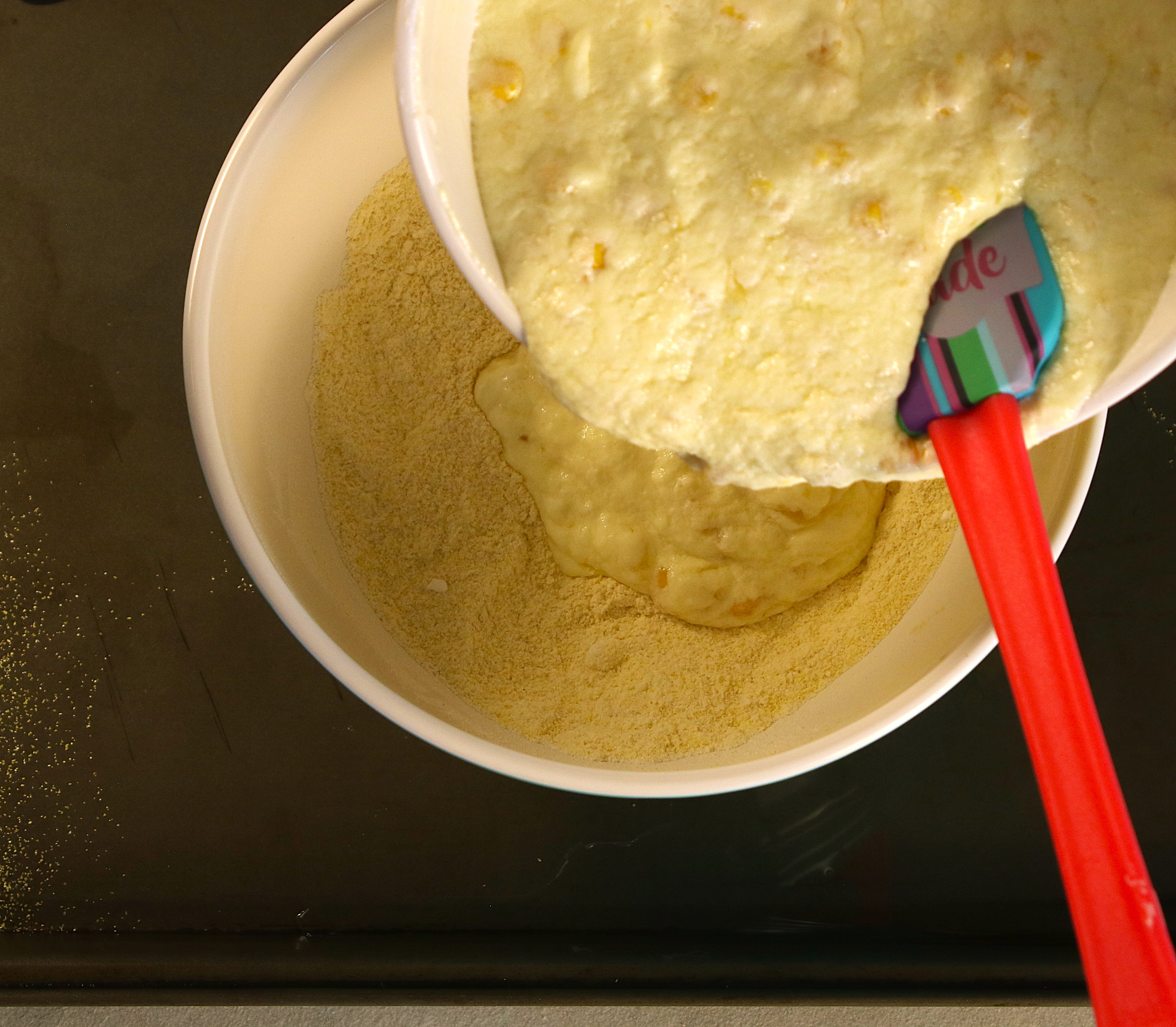 Wet mixture with corn being poured into the bowl of dry cornmeal mixture with a spatula.
