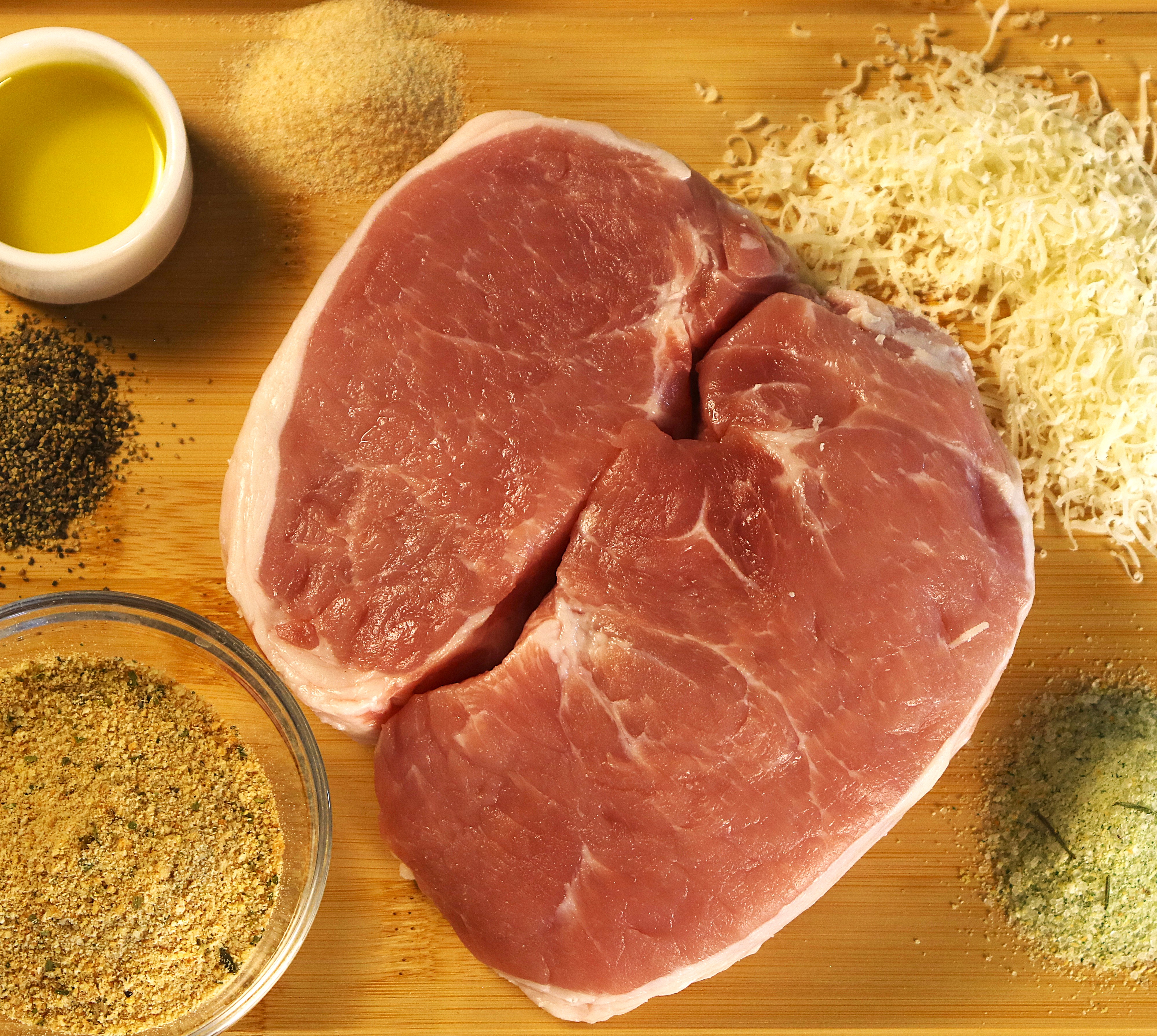 Overhead view of ingredients for parmesan breaded pork chops displayed on a wooden board, including two raw boneless pork chops, shredded parmesan cheese, Italian seasoned bread crumbs, olive oil, garlic salt, coarse black pepper, and onion powder.