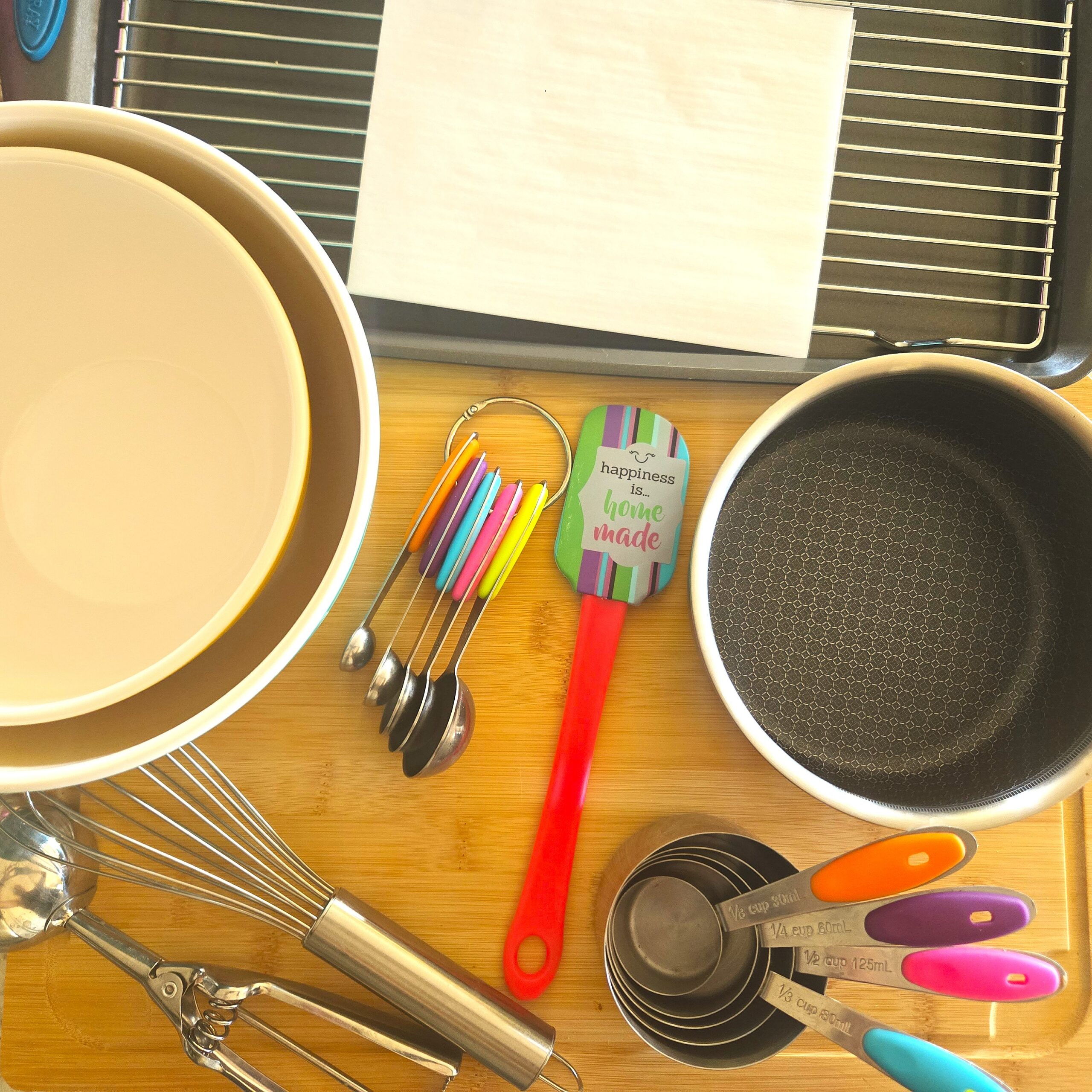 Overhead view of the instruments used to make White Chocolate Raspberry Cheesecake Cookies, including mixing bowls, a whisk, cookie scoop, measuring cups and spoons, silicone spatula, saucepan, baking sheet with parchment paper, and cooling rack arranged on a bamboo board.