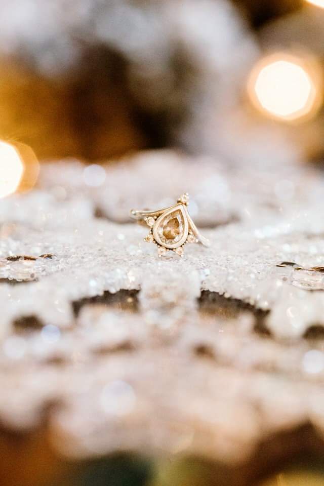 The Importance of Detail Photos: Rings, Florals & Personal Touches by GSquared Weddings Photography Seattle Snohomish Snohomish Wedding Photographer GSquared Weddings A gold ring with a teardrop-shaped setting is nestled on a detailed, sparkling snowflake ornament. The background is softly blurred with warm, glowing lights, enhancing the festive atmosphere. Serving Seattle, Snohomish and Orlando