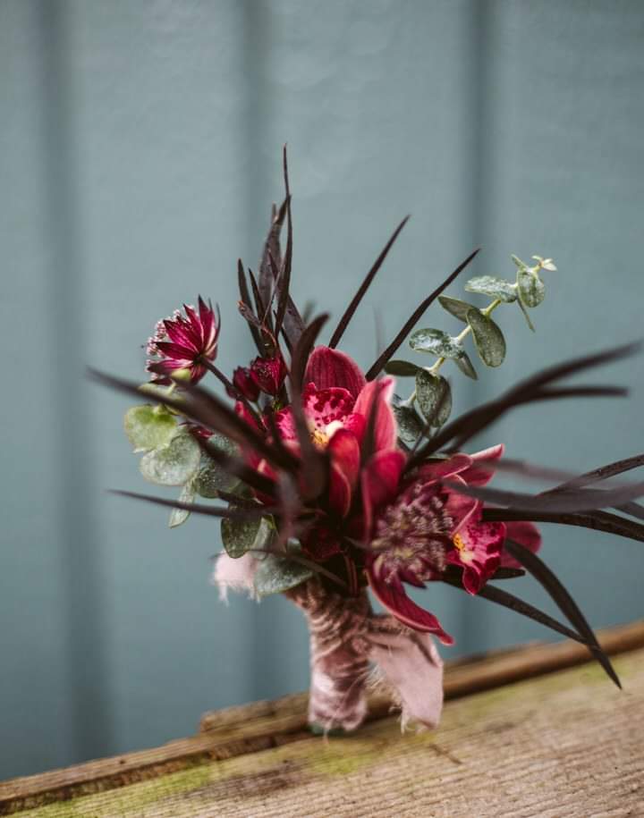 The Importance of Detail Photos: Rings, Florals & Personal Touches by GSquared Weddings Photography Seattle Snohomish Snohomish Wedding Photographer GSquared Weddings A small bouquet featuring red flowers, dark green leaves, and eucalyptus, wrapped in twine, rests on a wooden surface with a blurred, teal-colored background. Serving Seattle, Snohomish and Orlando