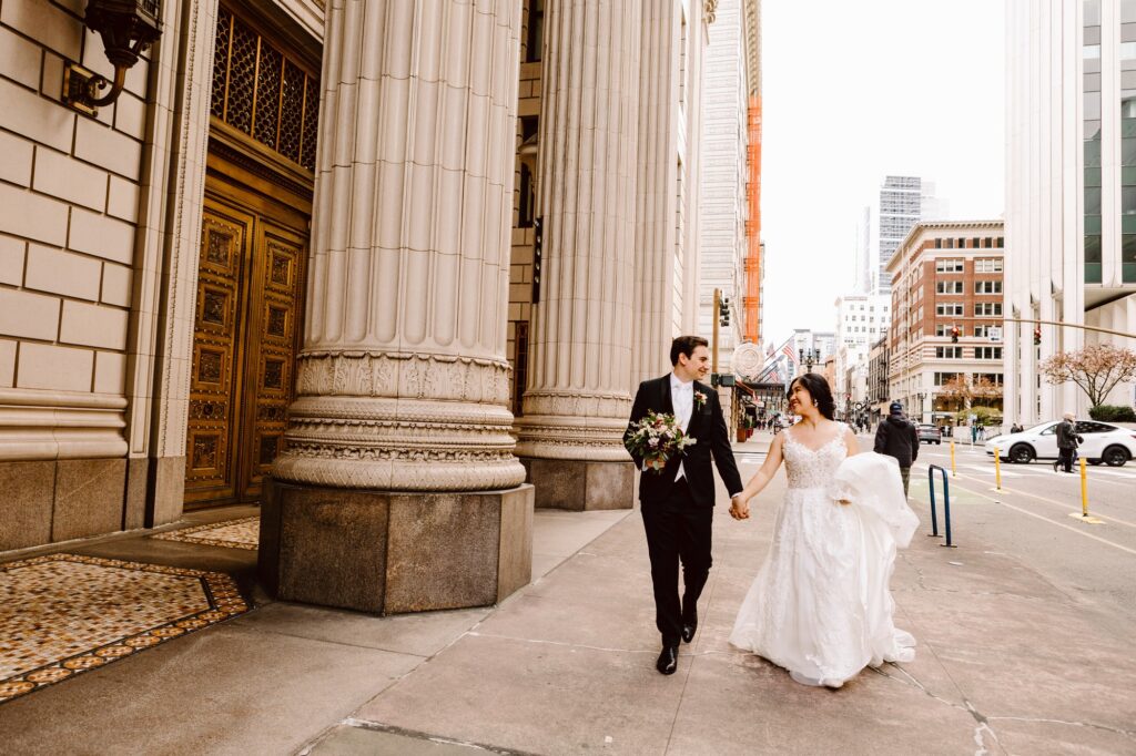 Snohomish Wedding Photographer GSquared Weddings A bride and groom joyfully walk hand in hand along a city street with tall buildings, taking a moment to pose. The bride wears a white gown, and the groom is in a black suit. Theyre smiling at each other, with the urban skyline visible in the background. Serving Seattle, Snohomish and Orlando