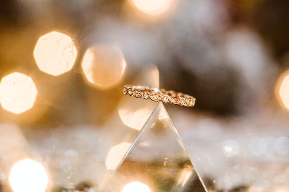 The Importance of Detail Photos: Rings, Florals & Personal Touches by GSquared Weddings Photography Seattle Snohomish Snohomish Wedding Photographer GSquared Weddings A gold ring with exquisite detail is displayed on a glass pyramid. The background features soft, glowing bokeh lights that create a warm, sparkling ambiance. Serving Seattle, Snohomish and Orlando