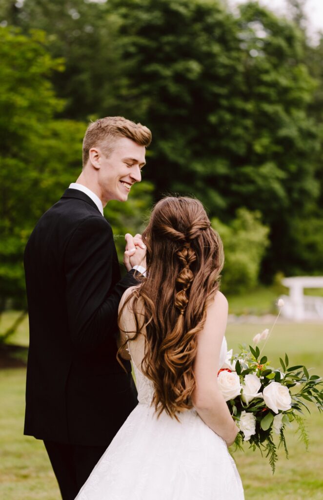 Snohomish Wedding Photographer GSquared Weddings A bride and groom stand outdoors, gracefully posing while holding hands. The groom wears a black suit, and the bride dons a white dress with her hair in loose waves. She cradles a bouquet of white flowers as lush greenery envelops them. Serving Seattle, Snohomish and Orlando