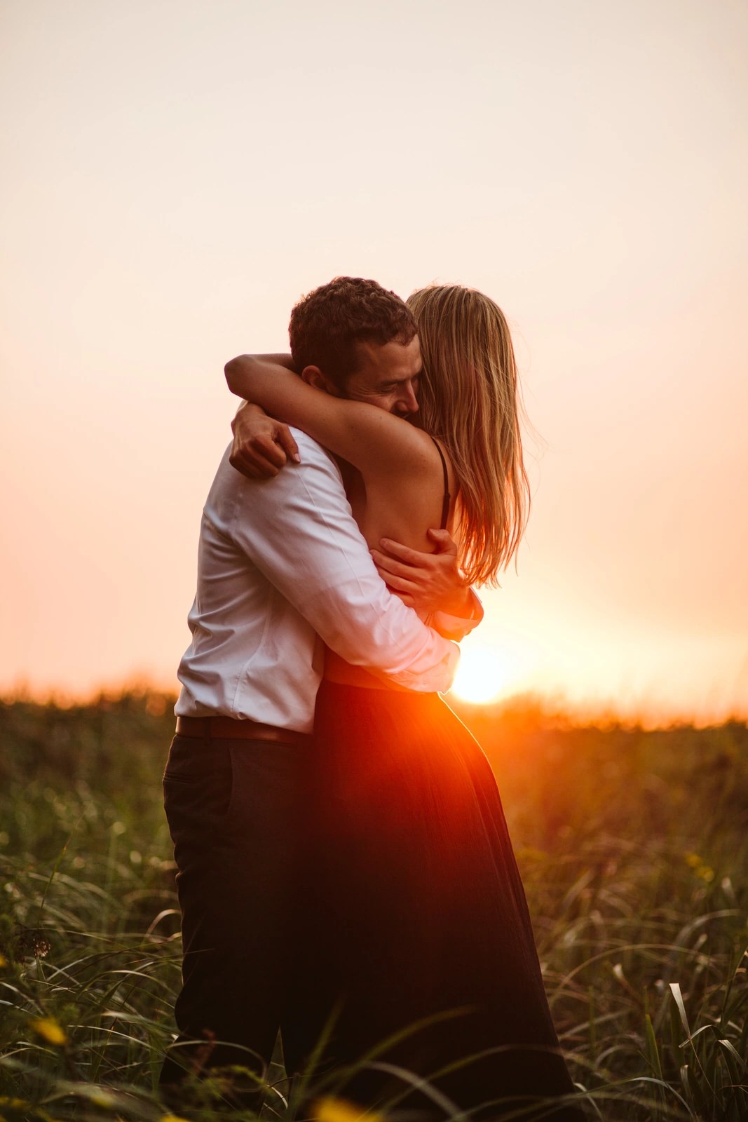 Snohomish Wedding Photographer GSquared Weddings A couple embraces in a grassy field at sunset, with warm sunlight glowing behind them. The woman wears a sleeveless dress, and the man wears a white shirt. They appear affectionate and peaceful in the natural setting. Serving Seattle, Snohomish and Orlando