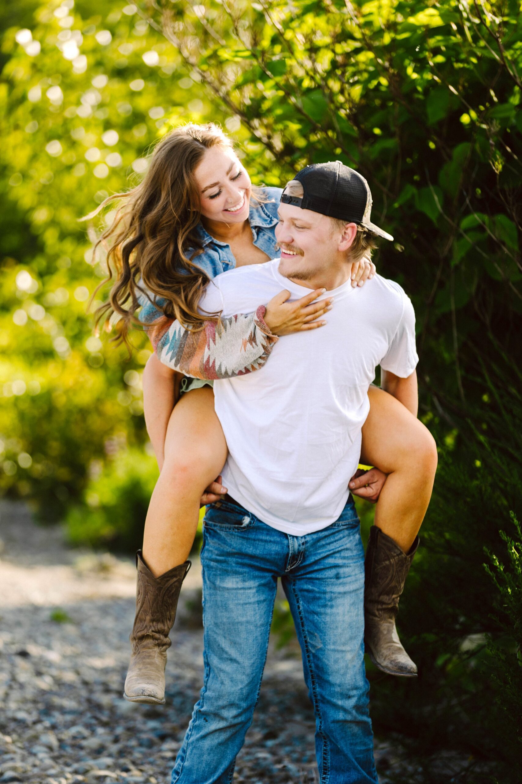 Snohomish Wedding Photographer GSquared Weddings A smiling man in a white t-shirt and jeans gives a piggyback ride to a laughing woman in boots and a denim jacket. They are outdoors on a sunny day surrounded by greenery. Serving Seattle, Snohomish and Orlando