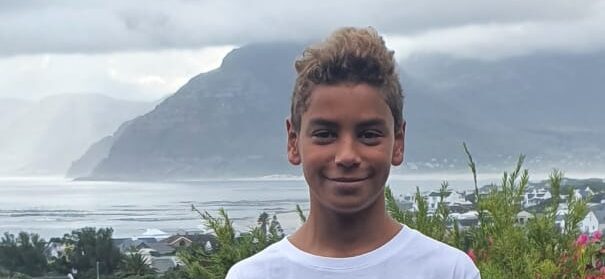 A portrait of a young man, smiling and wearing a white t-shirt, standing in front of a scenic coastal landscape featuring a sandy beach, houses, and mountains under a cloudy sky.