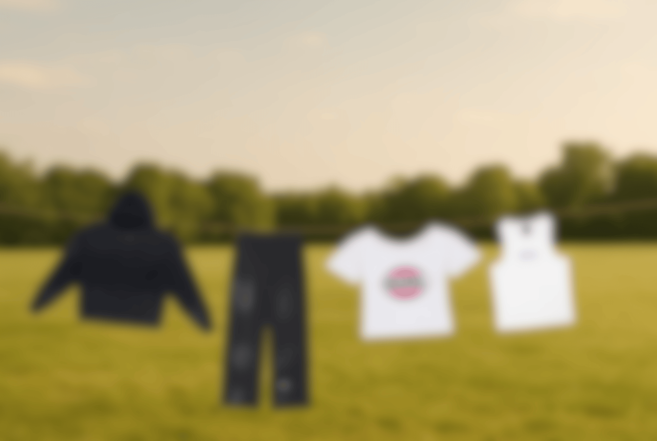 Four GURL clothing items — a black hoodie, black sweatpants, a white cropped tee with a pink-green logo, and a white halter tank — hanging on a clothesline in a wide green field at golden hour, softly blurred for a calm, natural, grounded mood.