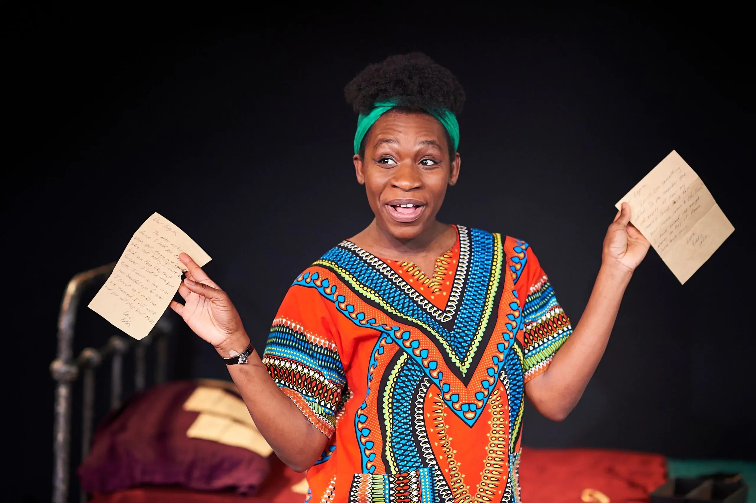 Apphia Campbell, a black woman wearing traditional kente clothing and a green headband, poses with two sheets of paper in her performance of Nina Simone