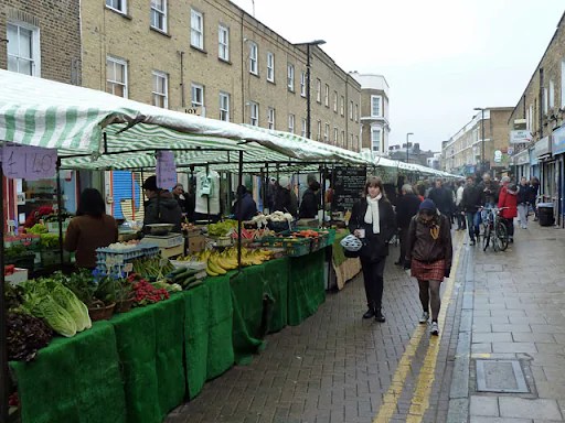 A row of market stalls, with green tablecloths that reach the ground, are in a row from right to the vanishing point of the center of the photo. It's a rainy day, but there are some people walking infront of the stalls, and the tables have colourful vegetables on them.