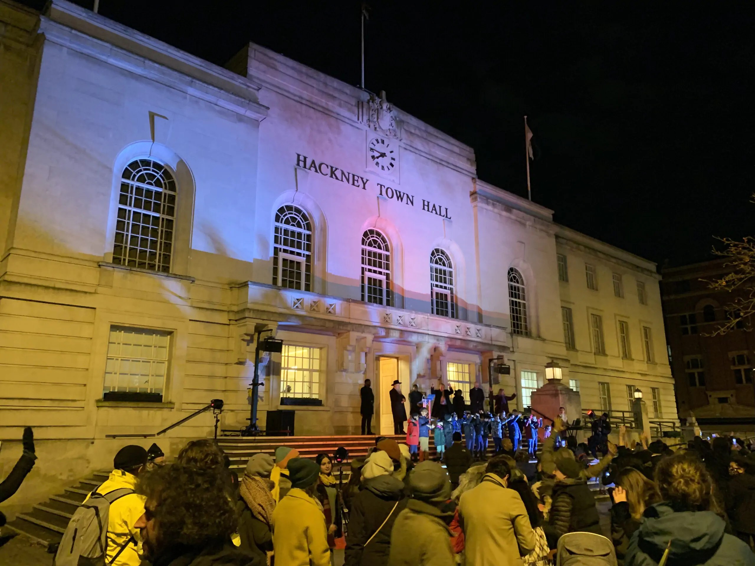 Descriptive photo of the Menorah lighting event. People gathered outside Hackney Town Hall to watch children sing Hanukkah songs.