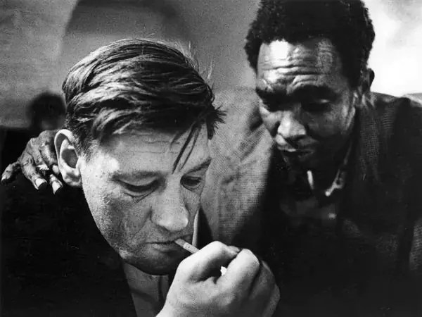 A black and white photo of a white man smoking, with a black man standing behind him with his hand on his shoulder.