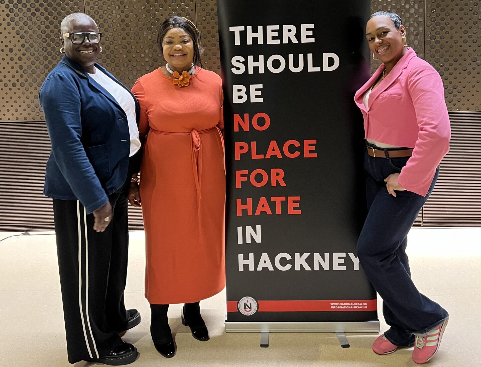 Three women are stood by a black banner which says: "#NationalHCAW, There should be no place for hate in Hackney" in large text