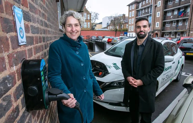 Mayor of Hackney Caroline Woodley, dressed in an indigo overcoat, stands next to an EV charger holding the nozzle.