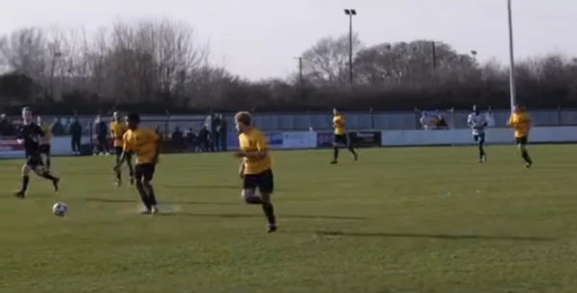 Hackney Wick during a counter attack, playing the ball forward into the opponent's half. Their kit is a bright yellow shirt with black shorts.