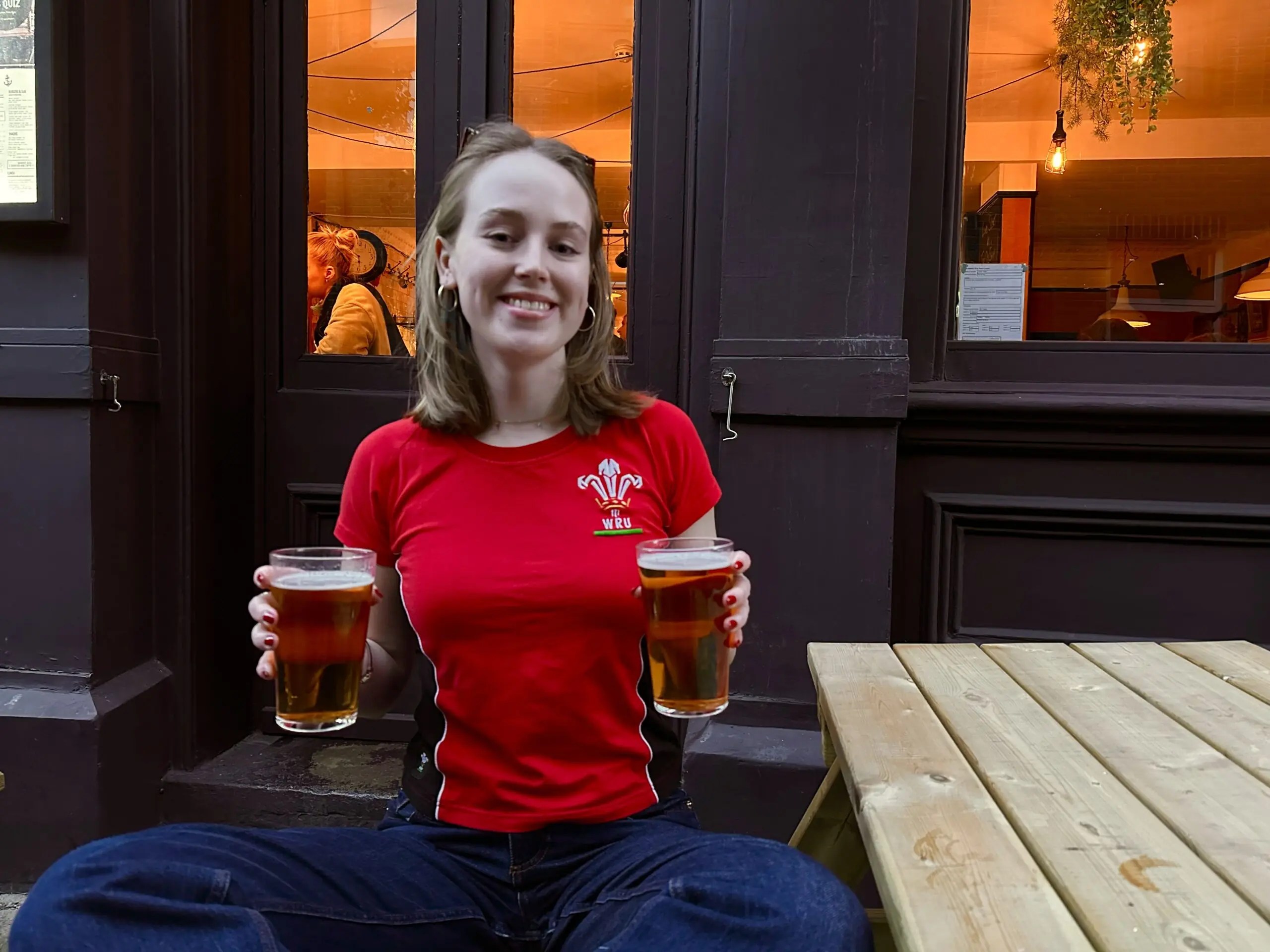 Our reporter Lily Scaplehorn (blonde hair)wearing a red Wales Rugby top smiles and poses with two full pint glasses of beer in her hands.