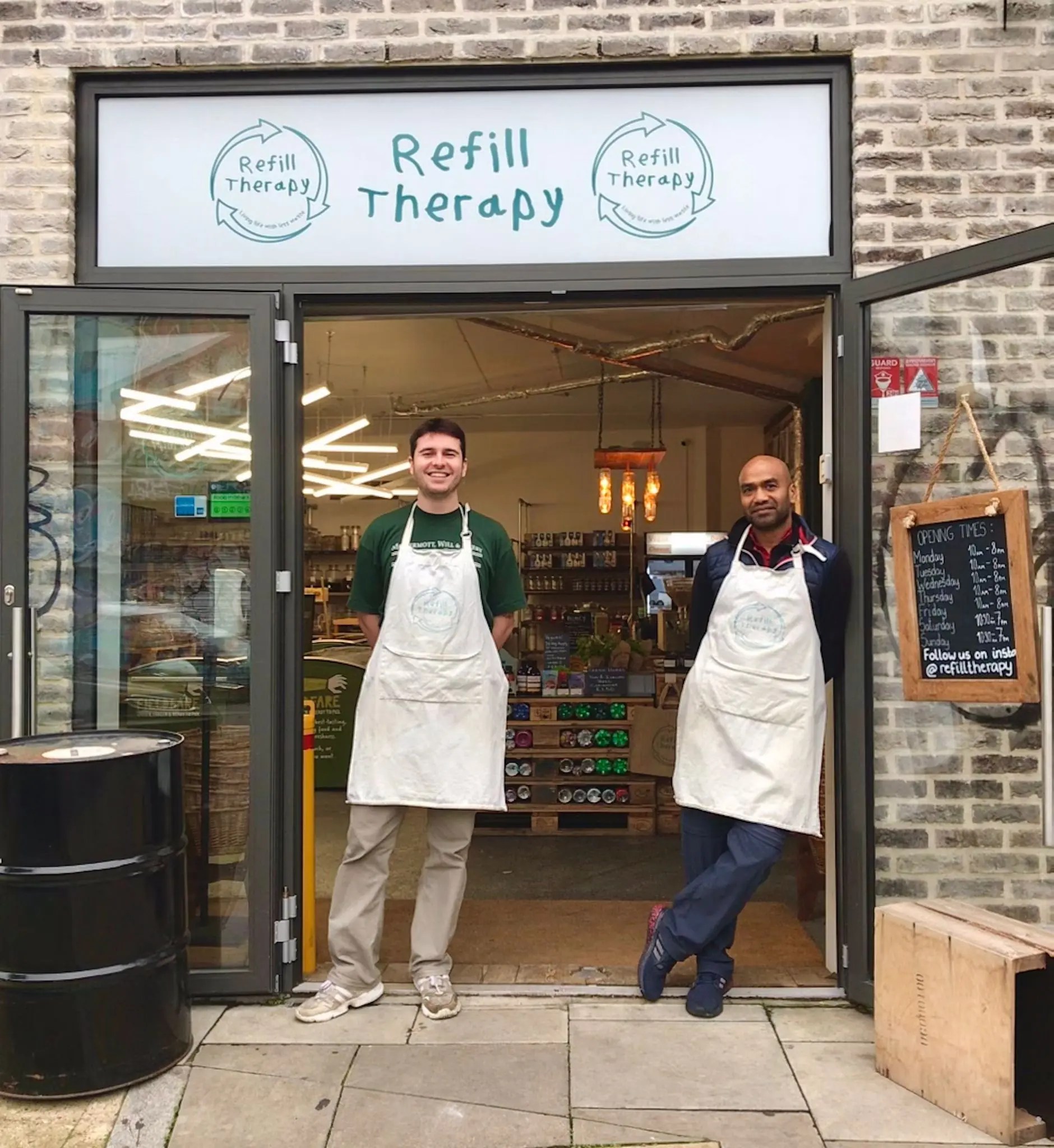 Two men wearing white aprons stand outside storefront