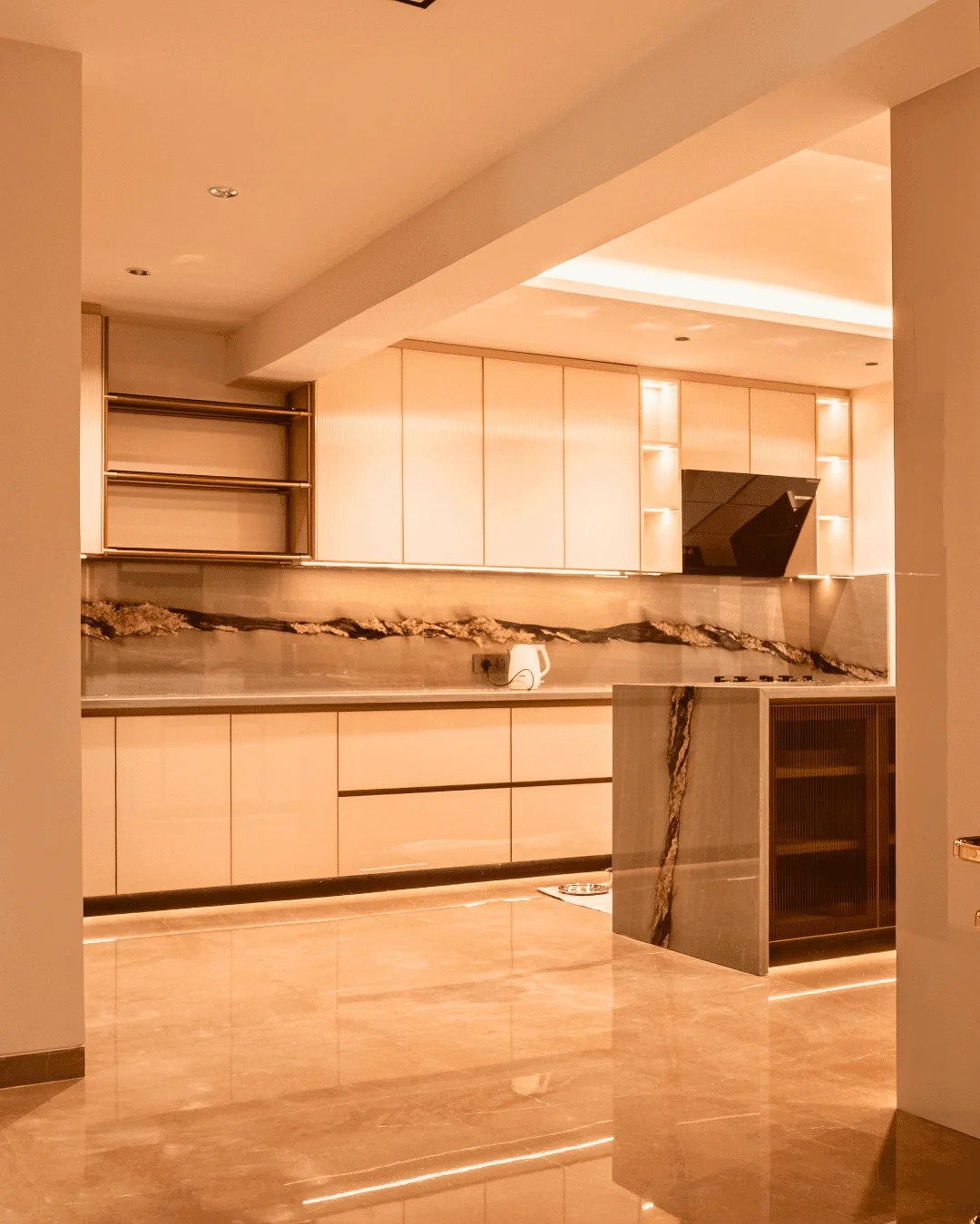 Sophisticated kitchen interior at GVK Skycity C301 showcasing glossy lacquered glass cabinetry, an island with dramatic veining, a matching marble backsplash, and a polished marble floor reflecting warm ambient light.
