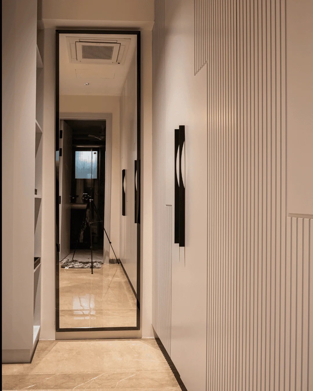 An architectural walk-in closet hallway showcasing seamless minimalist cabinetry with a pristine lacquered finish. The design features elegant linear fluted textures, sleek dark vertical hardware, and a slim-profile full-length mirror, creating a refined perspective.