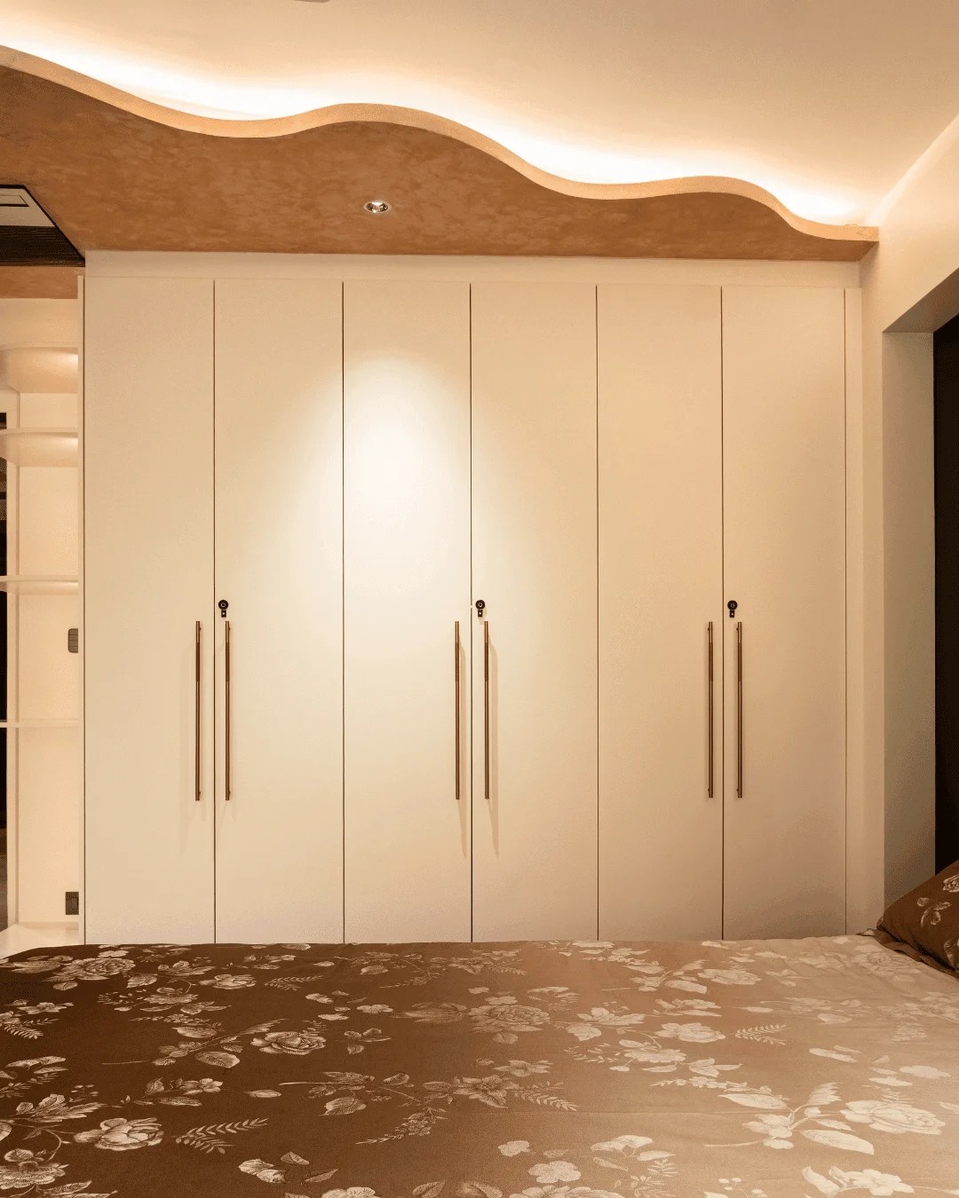 The master bedroom of a GVK private residence, highlighted by a floor-to-ceiling bespoke wardrobe with a clean, high-gloss lacquered finish. The space is crowned by an architectural wave false ceiling with warm, hidden LED profile lighting, defining a curated, serene luxury space.